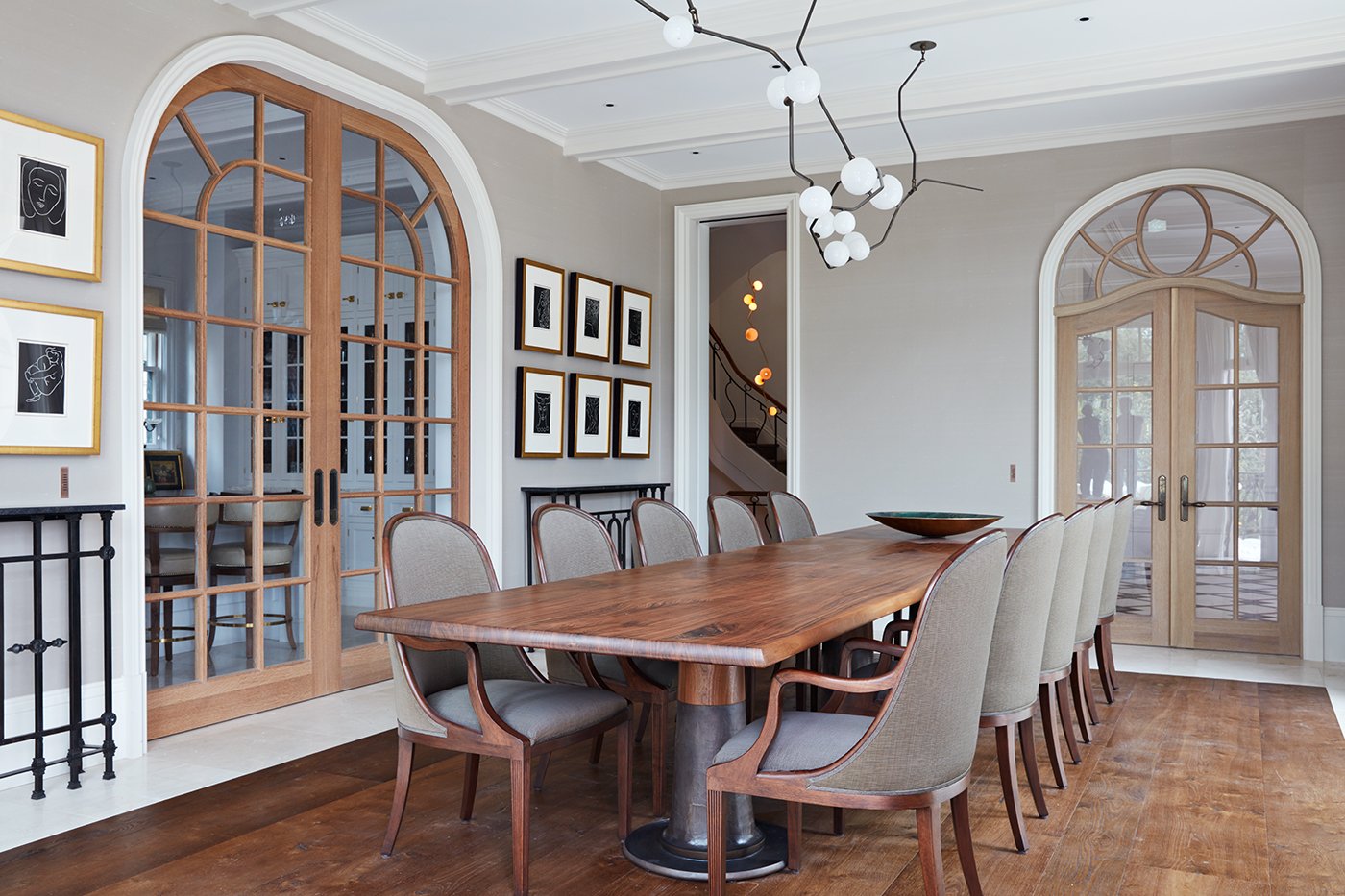 Elegant dining room with a large wooden table, beige upholstered chairs, modern chandelier, framed artwork on the wall, and glass double doors.