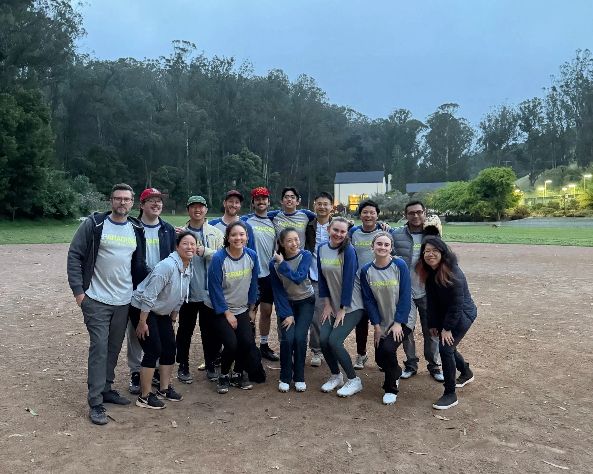 A group of young adults and teenagers standing outdoors on a dirt field, smiling and posing for the photo. They are dressed in sportswear, some wearing matching shirts with the word 'PARADIGM' on them. In the background, there are trees, a house, and