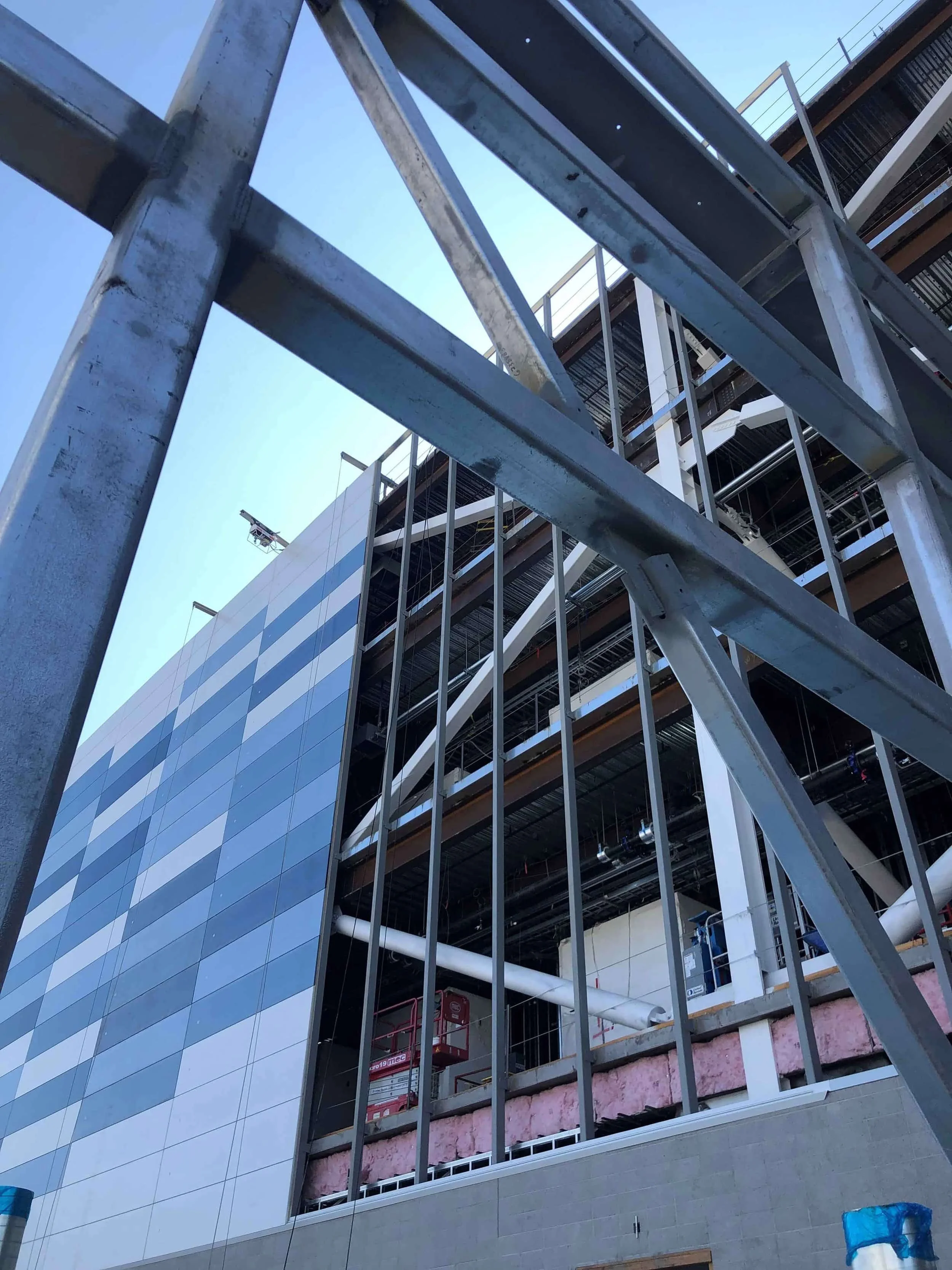 Construction site of a multi-story building with steel framing, insulation, and exterior glass panels, viewed from below through a metal fence.