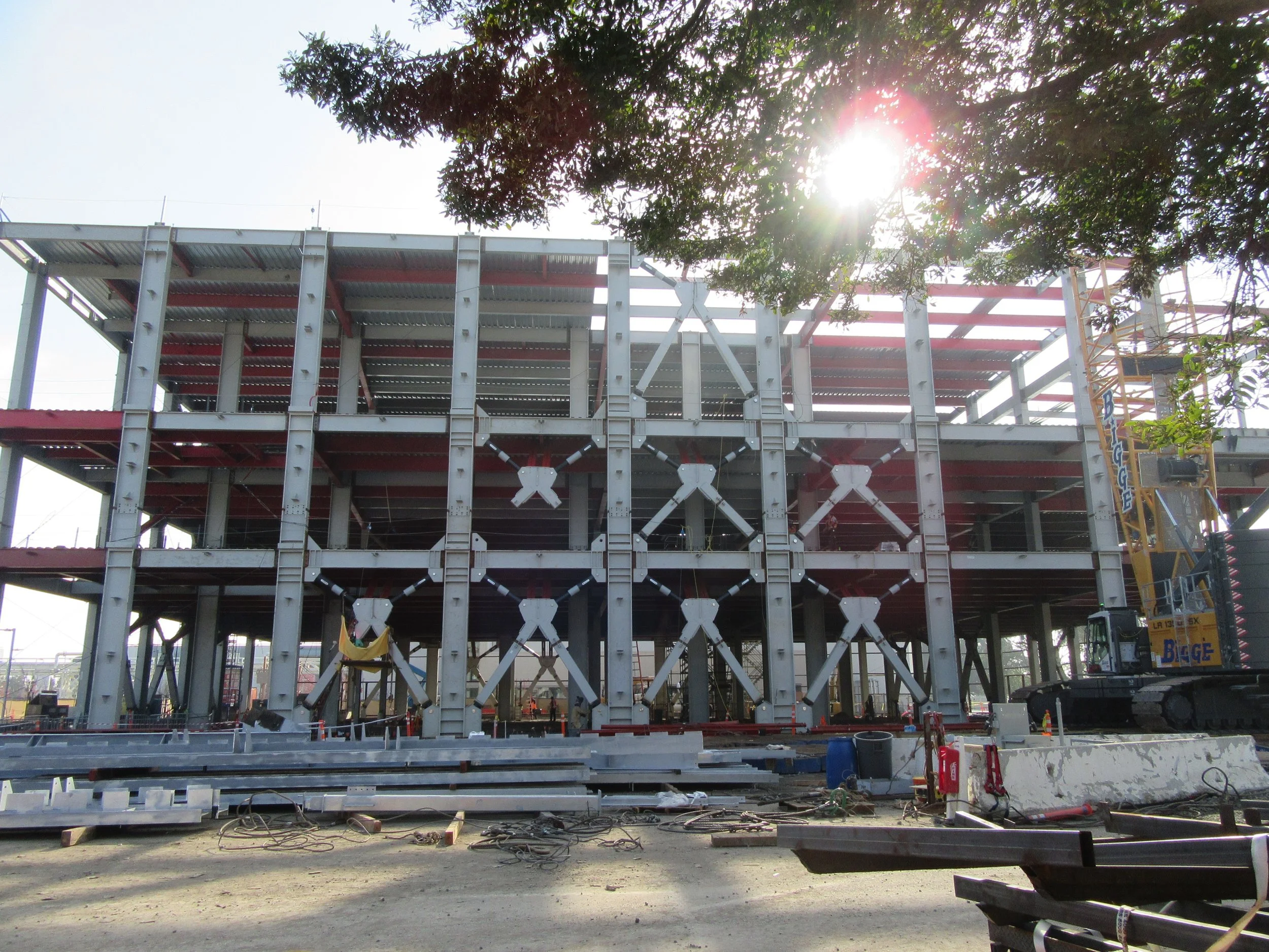Construction site with a multi-story steel framework, a crane on the right, and sunlight shining through tree branches at the top.