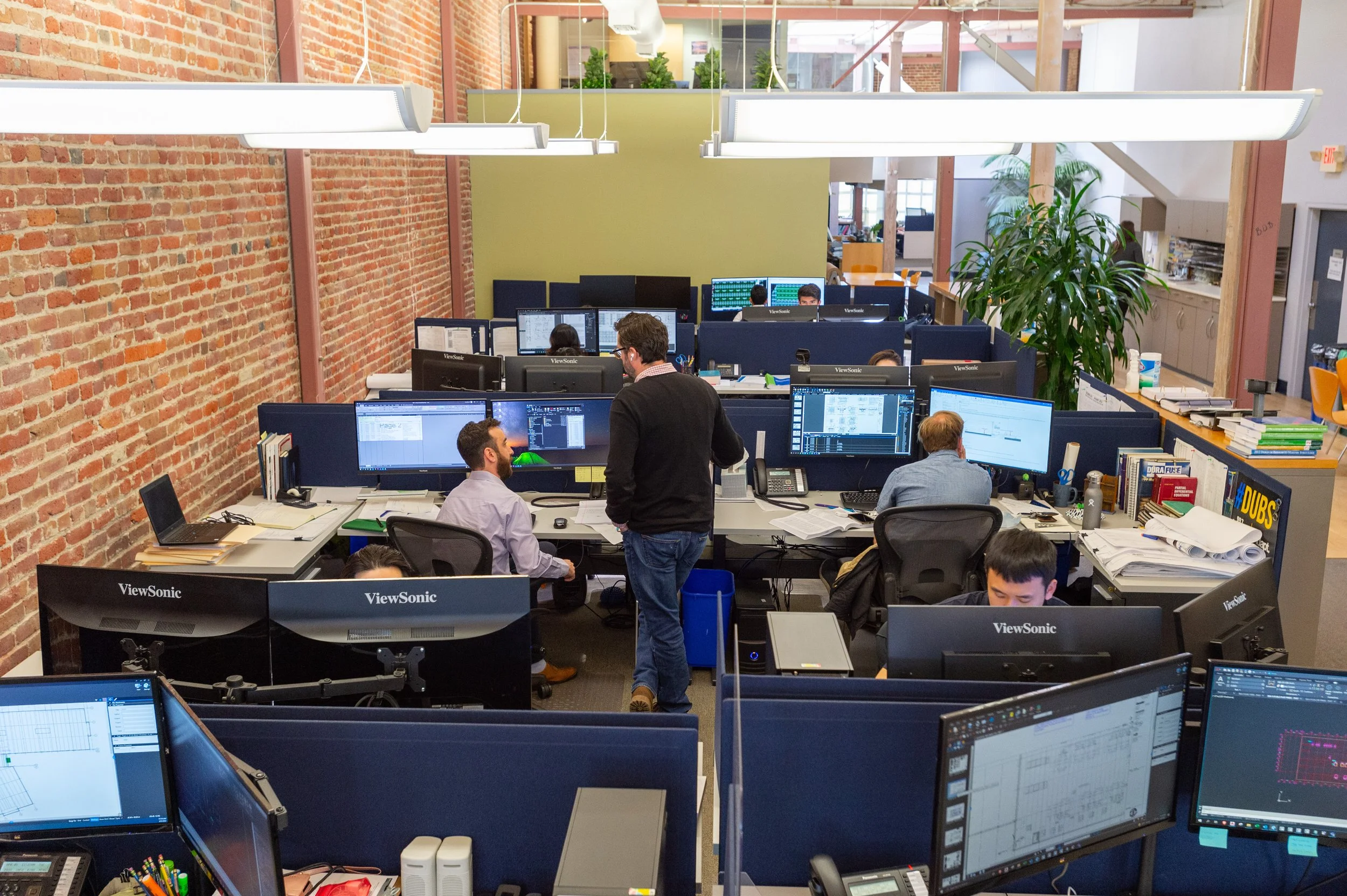 Open-plan office with multiple employees working at desks with computer monitors, brick wall on the left, and large windows in the background, with a large green plant and yellow chairs.
