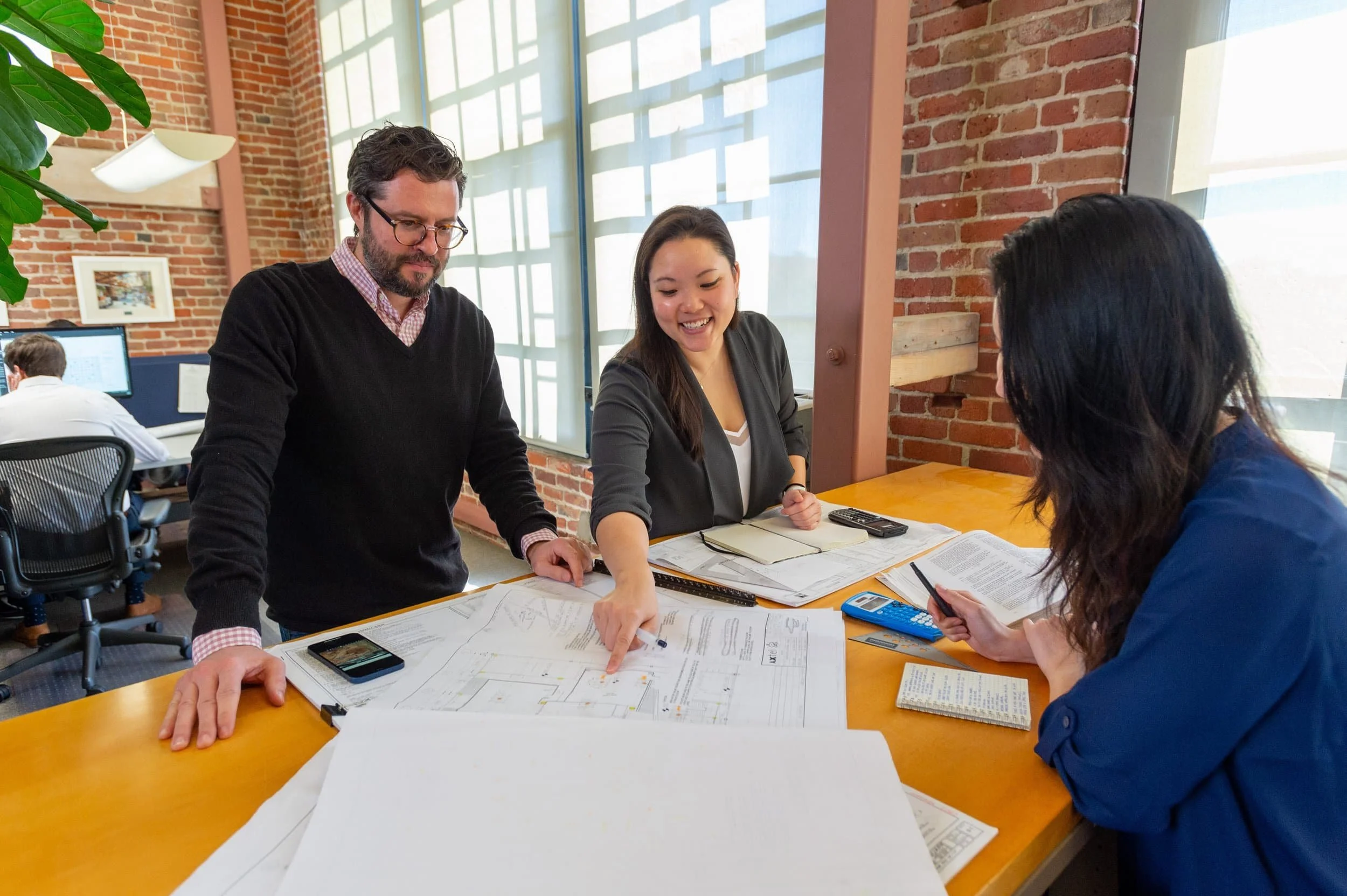 Three colleagues working together at a table in an office, examining architectural plans and documents, with a brick wall and large windows in the background.