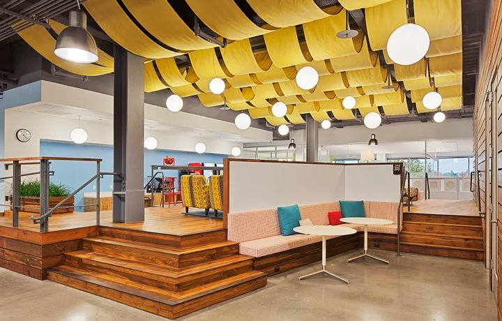 Interior office breakout space designed for PARADIGM Structural Engineers featuring multi-level wooden platforms, built-in booth seating with small tables, and a decorative yellow fabric ceiling canopy with pendant lights.