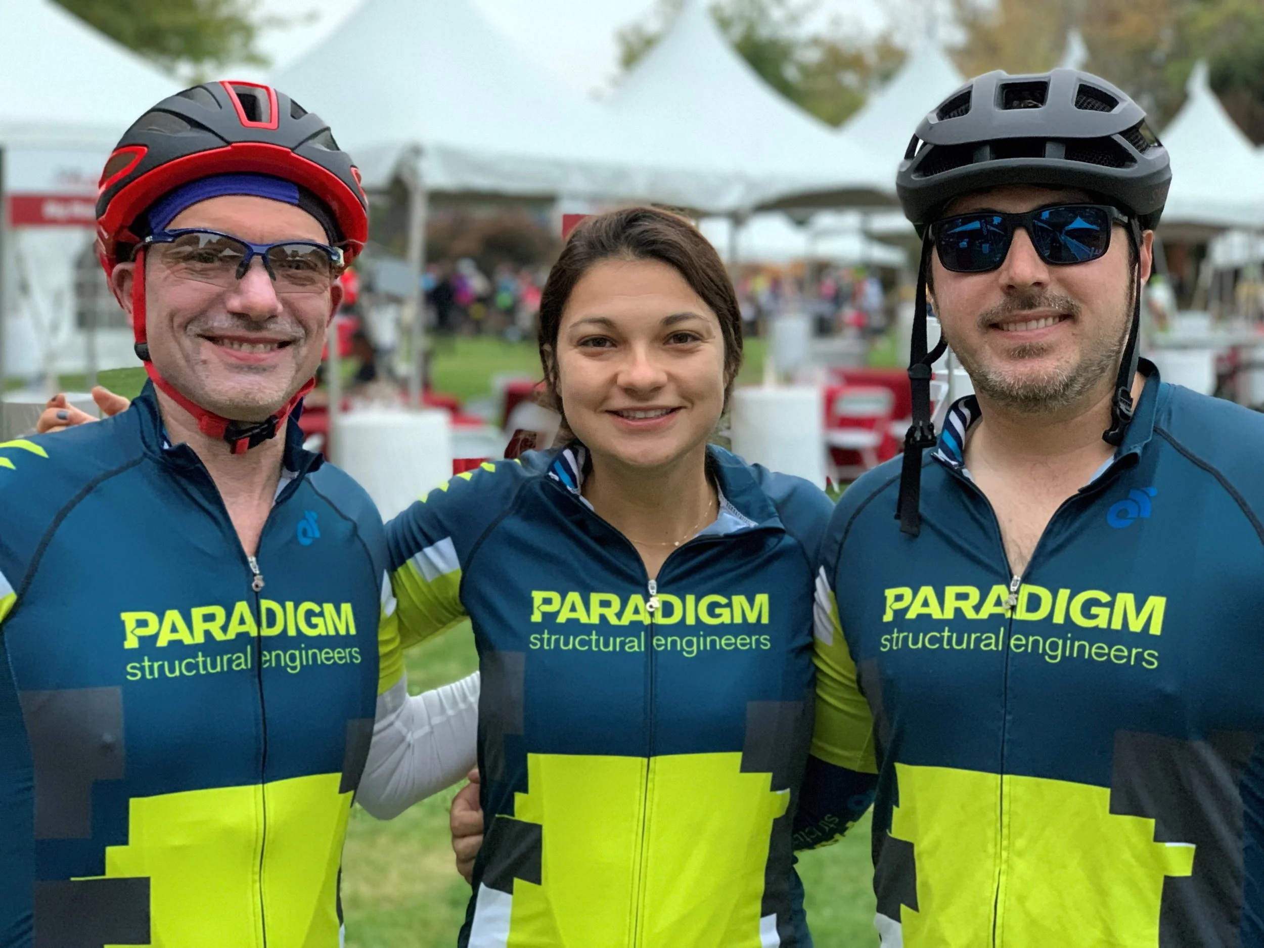 Three people in cycling jerseys standing outdoors, smiling, with tents and other people in the background.