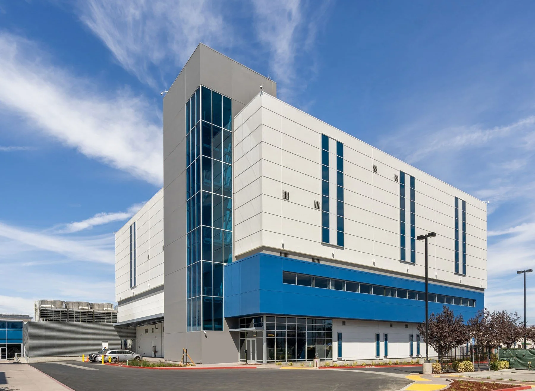 Daytime exterior view of the PARADIGM Structural Engineers Vantage CA15 data center facility featuring a multi-story facade, prominent blue and white panels, and a tall vertical glass tower under a clear blue sky.