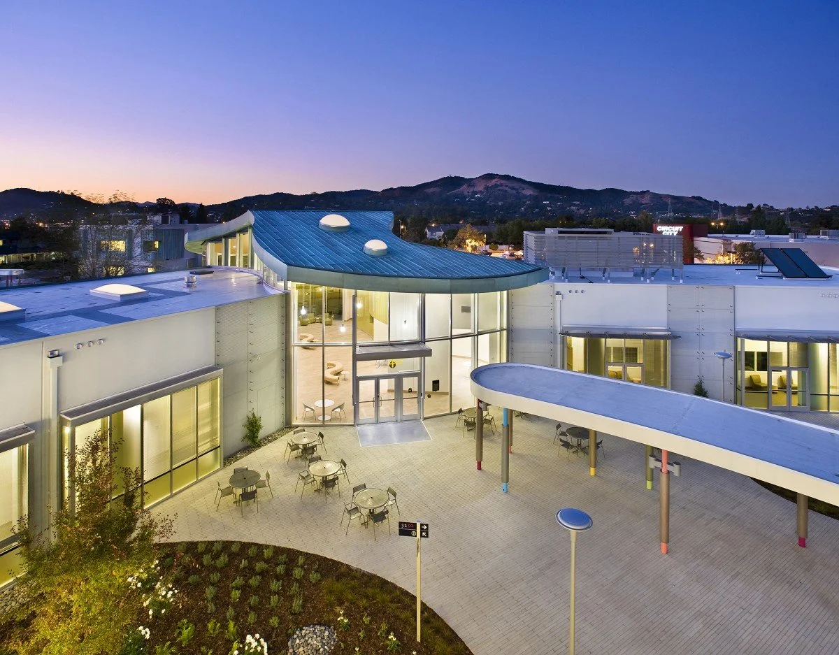 Modern building with large glass entrance, outdoor seating area, blue curved roof, and mountain background at dusk.