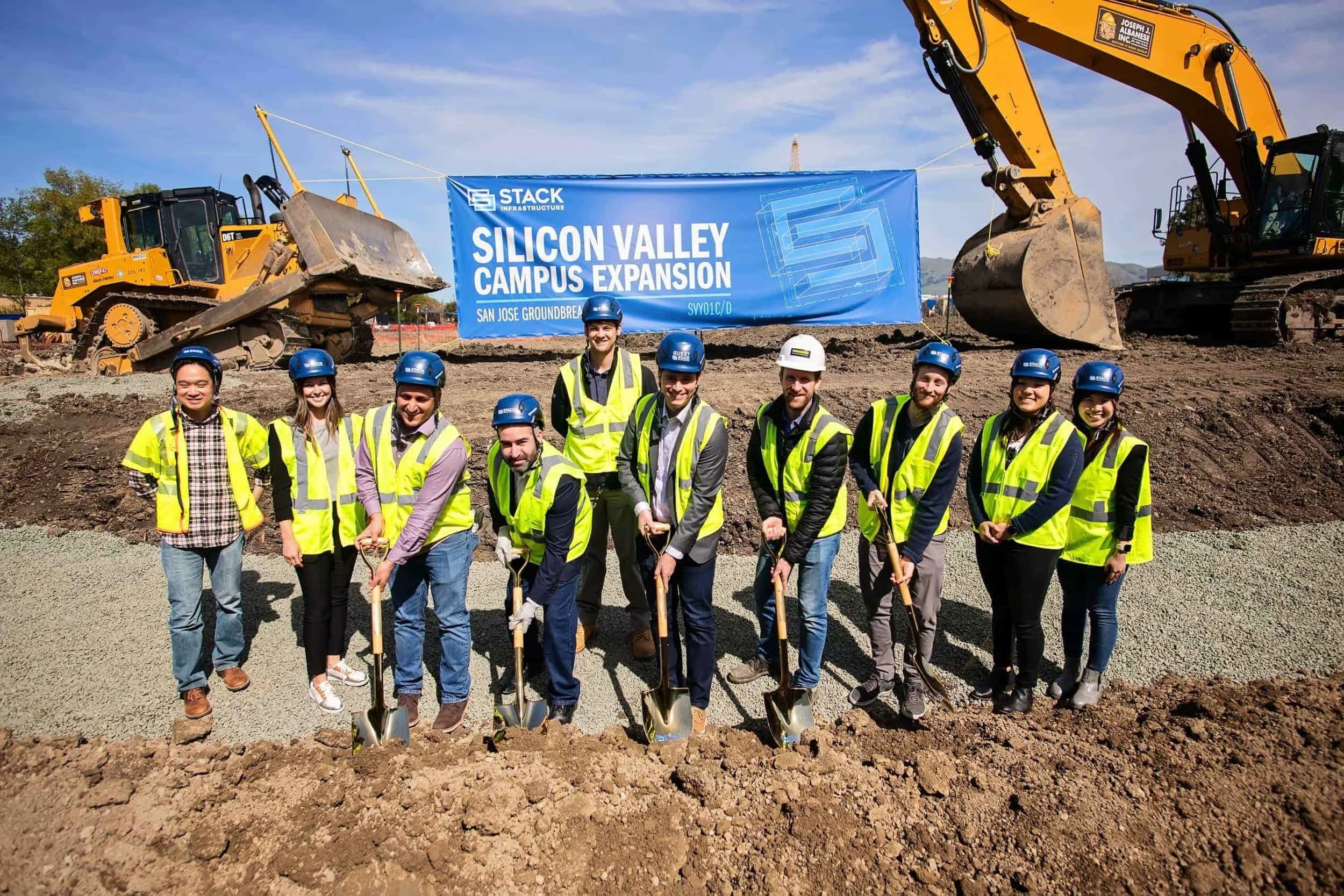 Group of people wearing yellow safety vests and helmets, holding shovels, at a construction site with heavy machinery and a banner that reads 'Silicon Valley Campus Expansion'.
