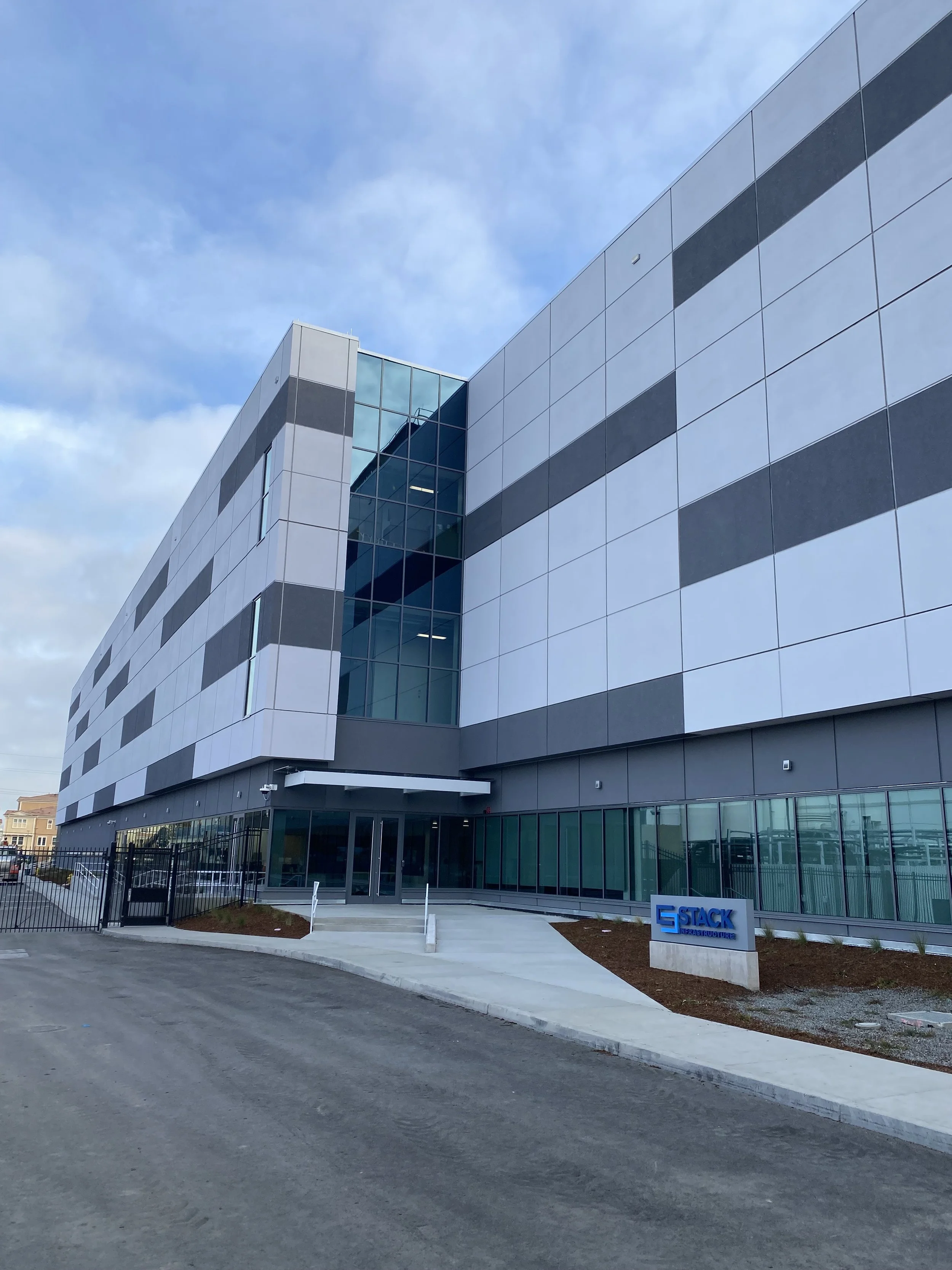 Exterior view of a modern office building with a glass entrance, gray and white panels, and a sign that reads 'STACK' near the sidewalk.