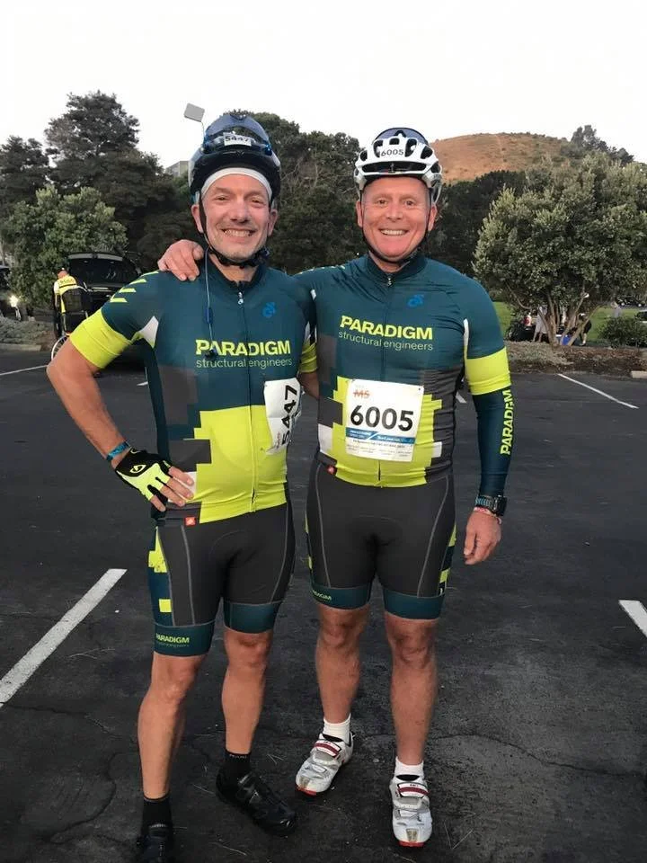 Two men in cycling gear standing together in a parking lot, smiling at the camera. They are wearing helmets and cycling uniforms with 'Paradigm structural engineers' written on them. One has a number 147, and the other has 6005 on their race bibs.