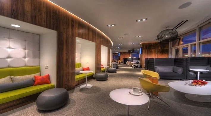 Modern lounge or hotel lobby with green and gray seating, small round tables, and large windows showing an evening city view.