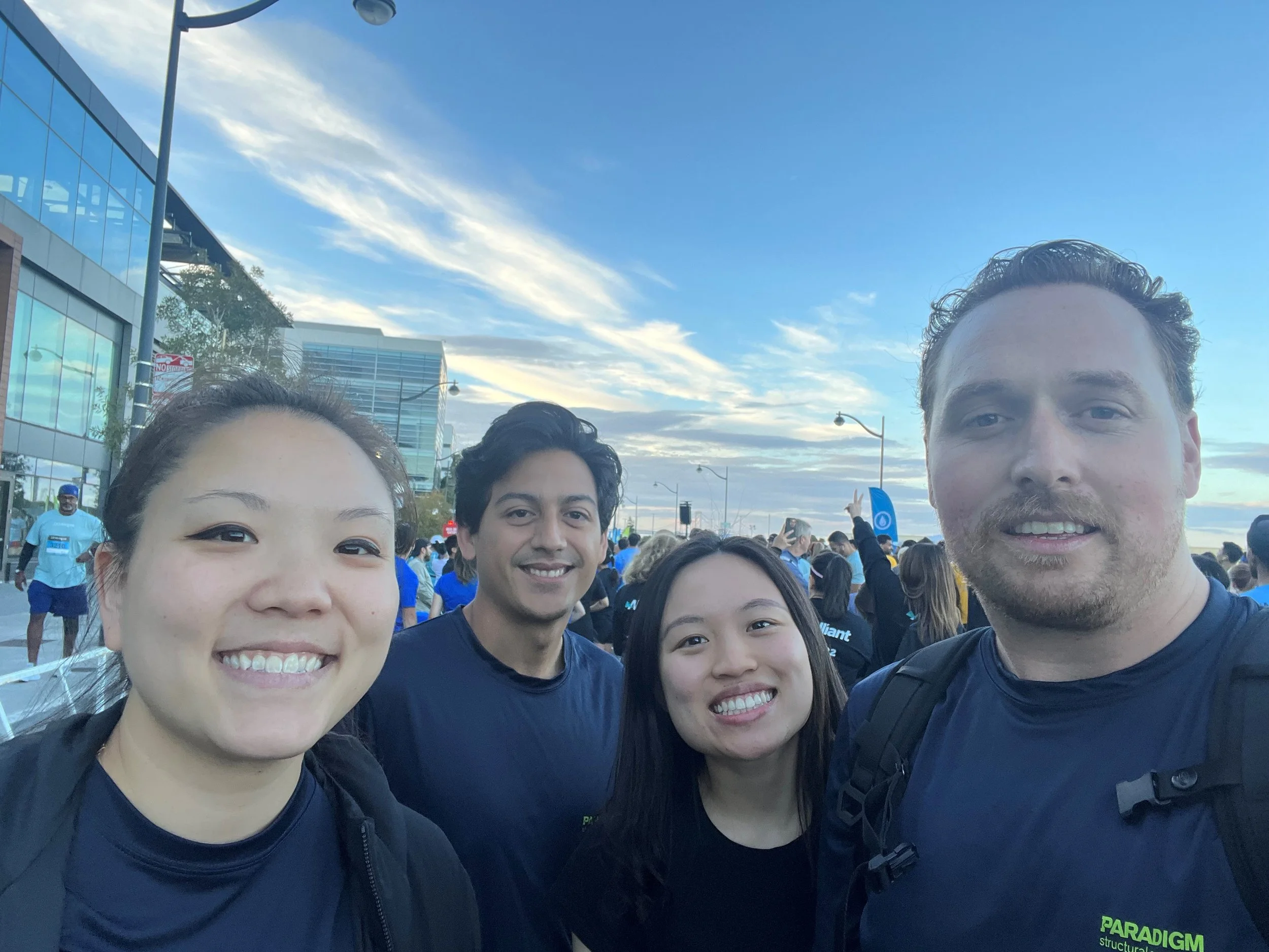 Four smiling people taking a selfie outdoors during a daytime event, with a crowd in the background and modern buildings under a partly cloudy sky.
