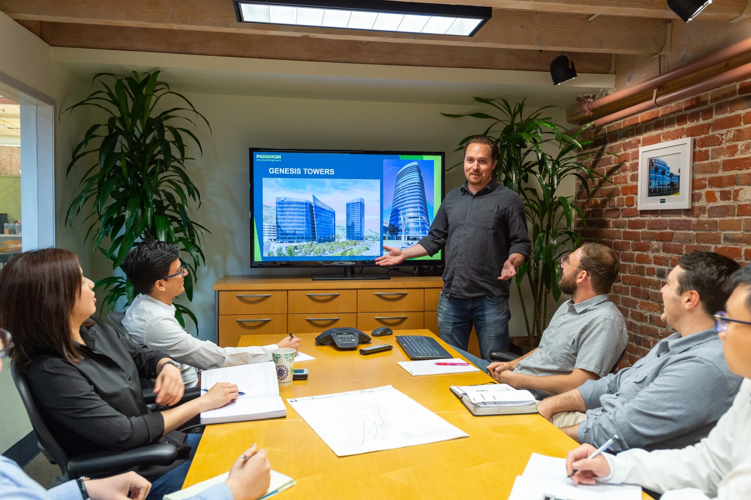 A man is giving a presentation to a group of six people in a conference room. The man is gesturing towards a screen displaying images of Genesis Towers. The room has a brick wall, tall plants, and a ceiling window.