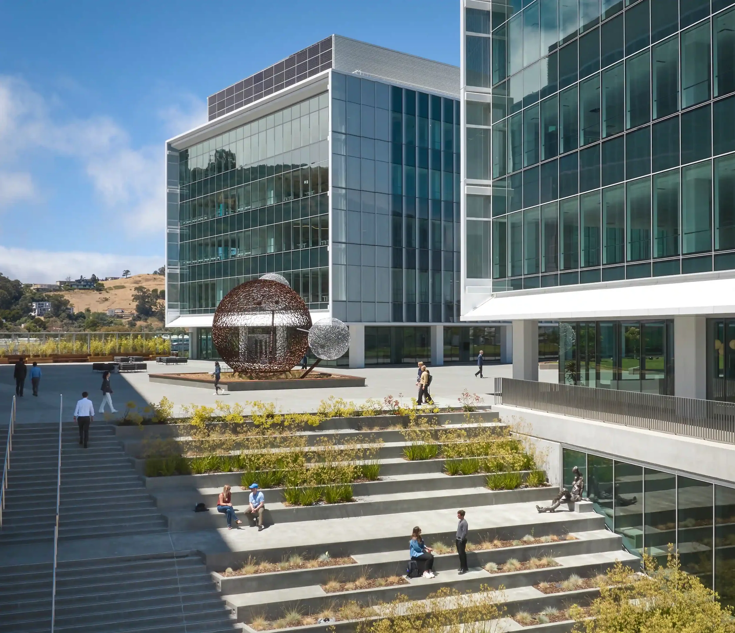 Modern office building with glass exterior, outdoor stairs with greenery, and abstract metal sculpture in front.