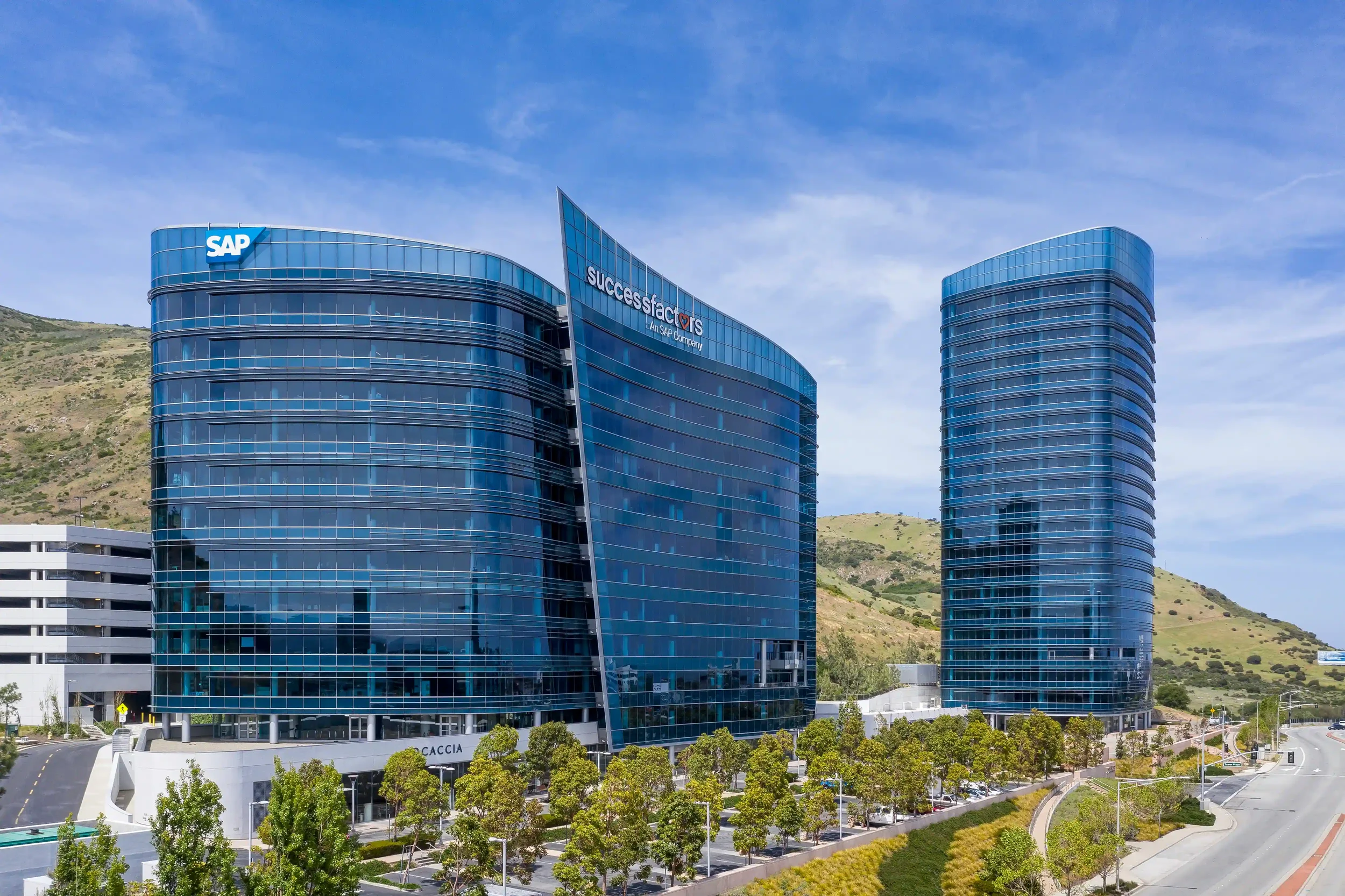 Two modern glass office buildings with SAP and SuccessFactors signage, surrounded by green trees and hills beneath a blue sky.