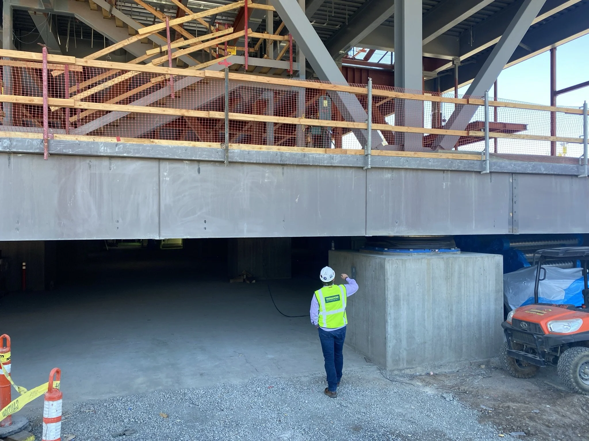 Construction site showing a worker in a safety vest and helmet walking under a partially built structure with scaffolding and wooden supports; a small utility vehicle parked nearby.