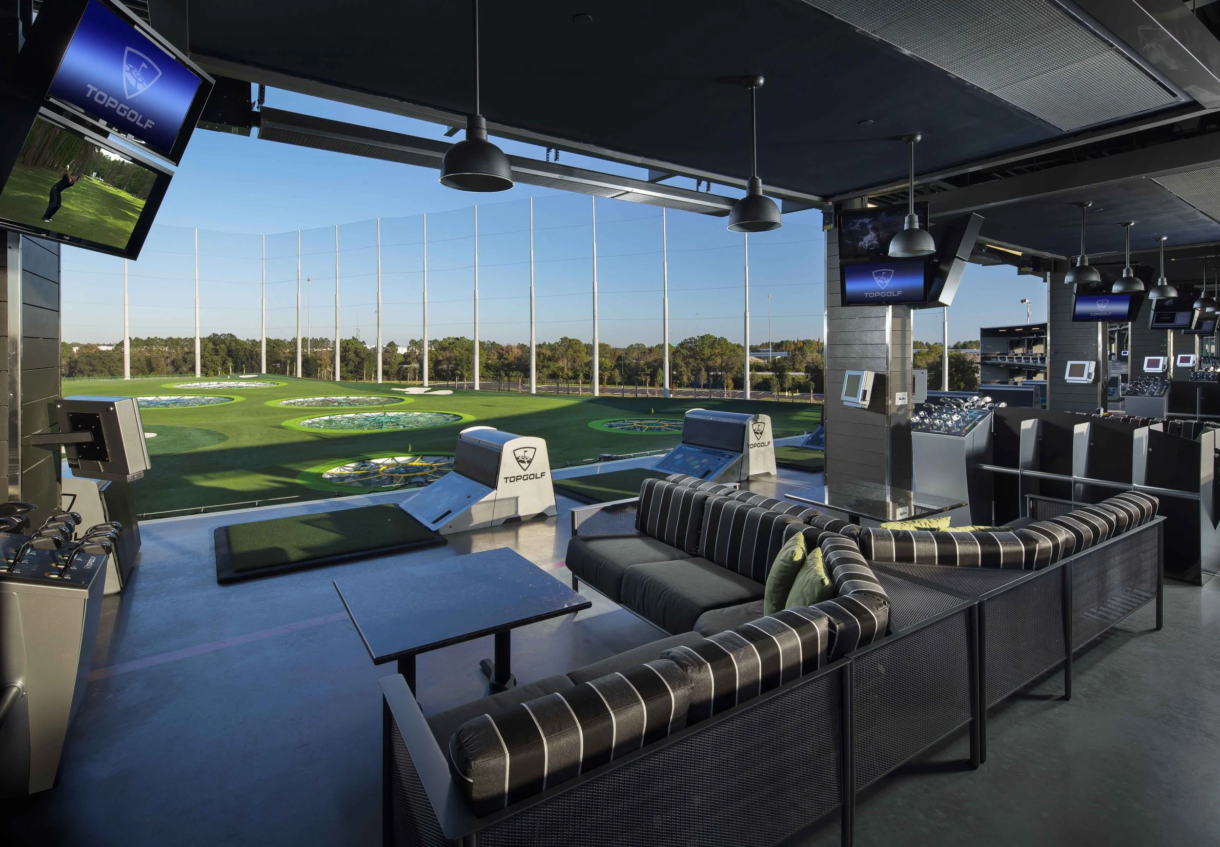 Indoor golf simulation lounge with multiple screens, a couch, and a view of outdoor golf mats.