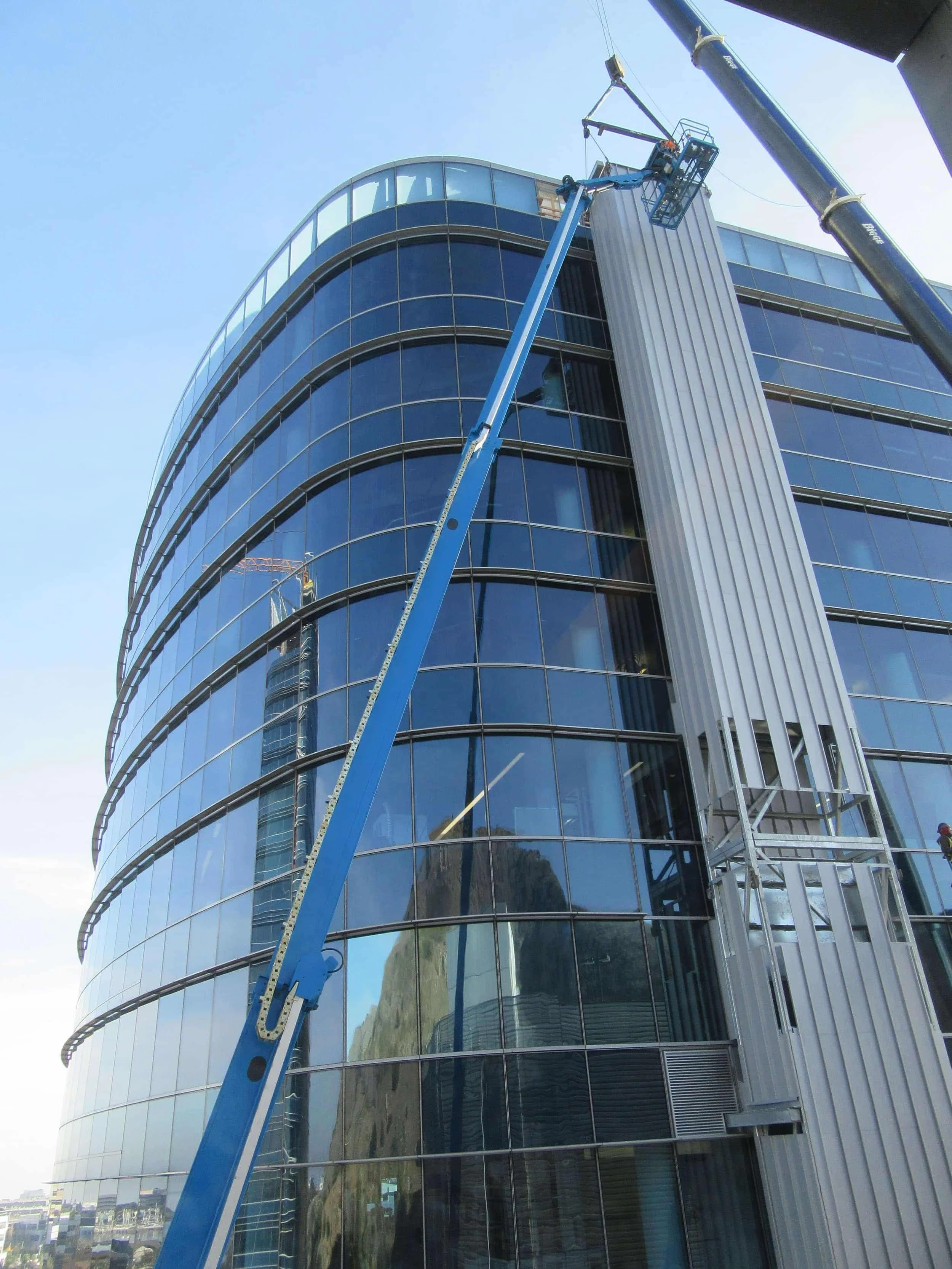 A curved glass building under construction with a blue crane and a worker on a lift.
