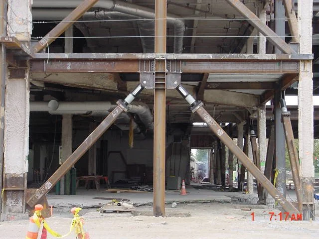 Construction site with steel support beams and scaffolding, with orange traffic cones and caution tape in the foreground.