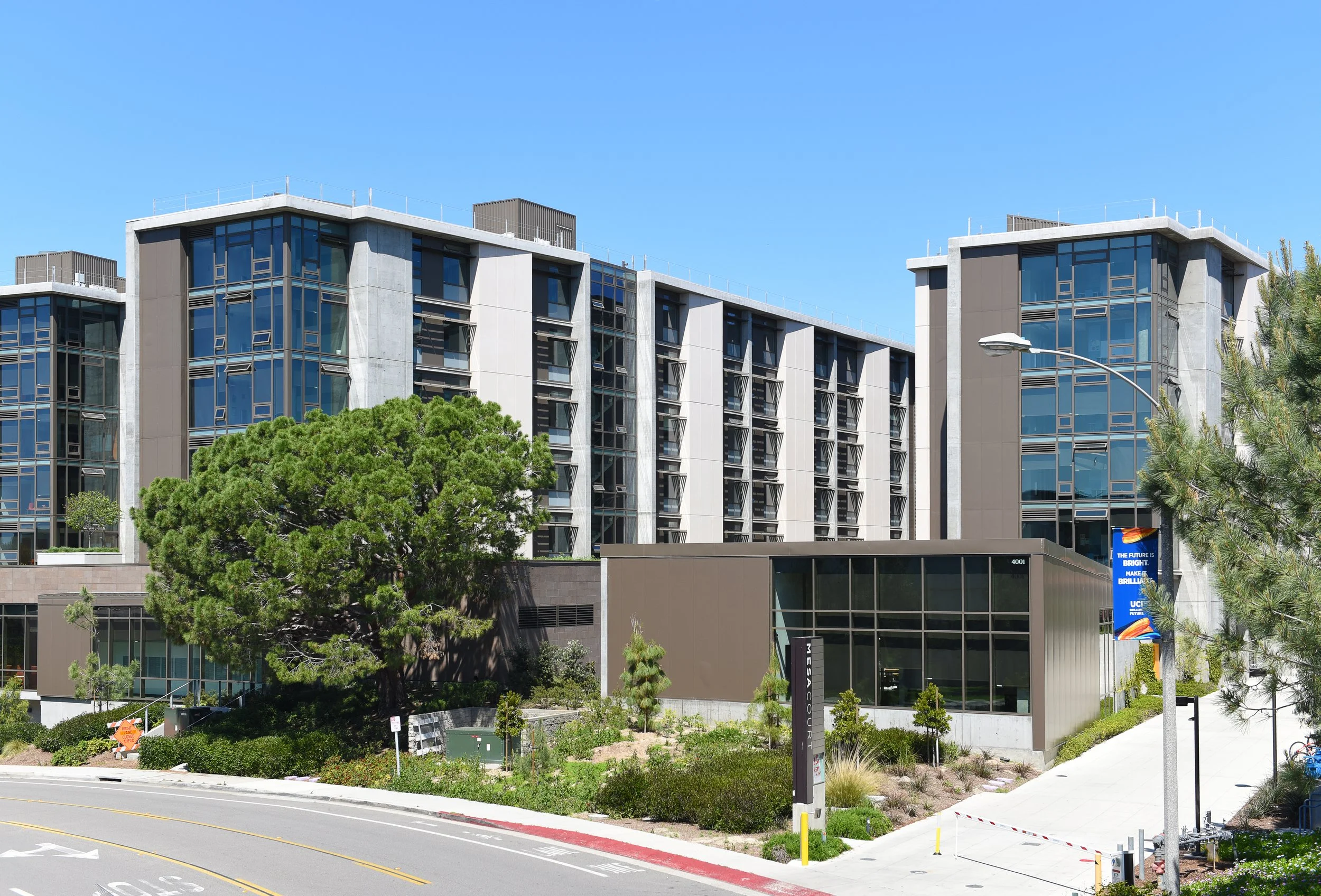 Modern multi-story university building with glass windows, greenery in front, and a blue banner on a street lamp.