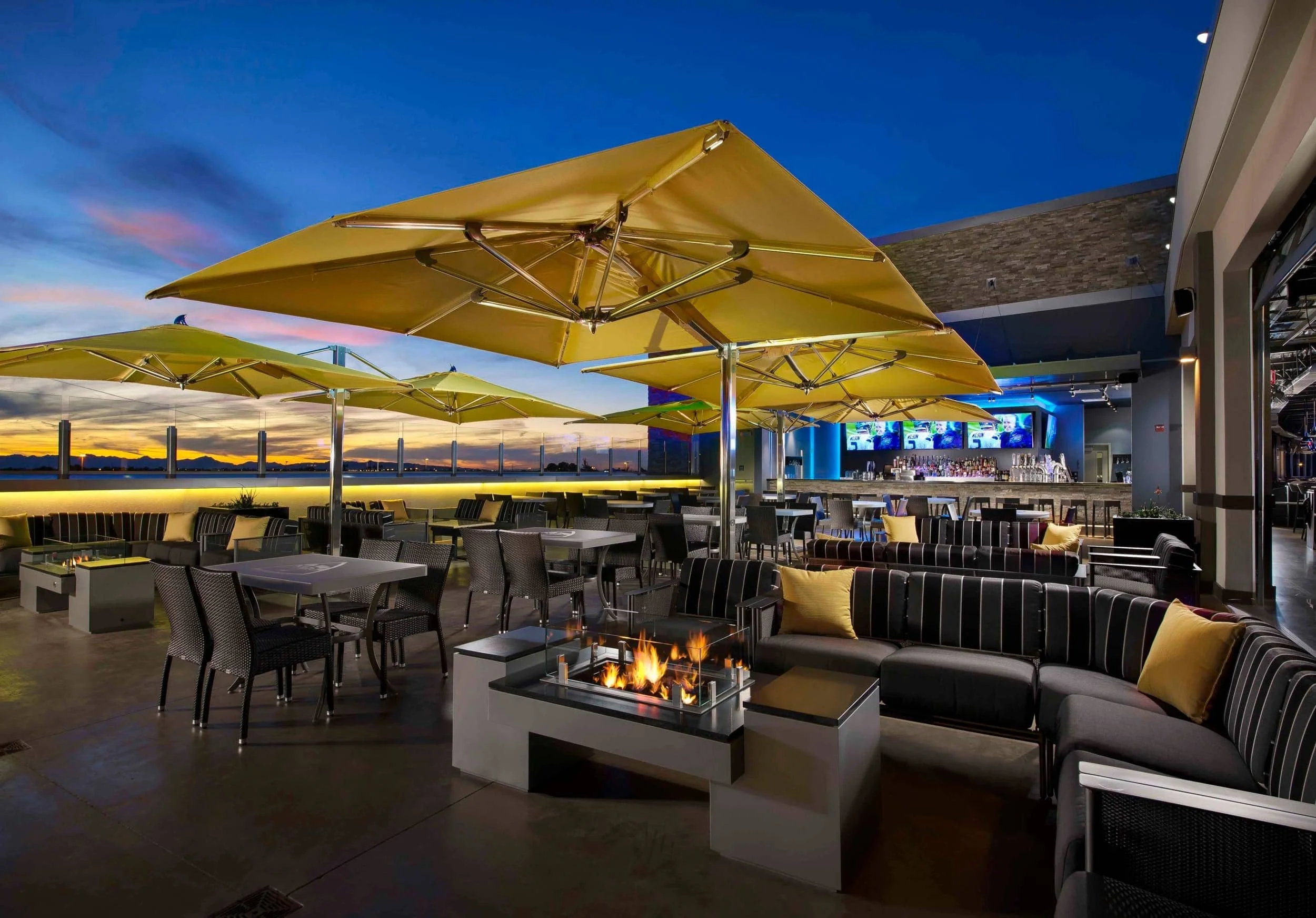 Twilight view of an outdoor rooftop lounge and bar patio featuring large yellow umbrellas, seating areas, and fire pits designed by PARADIGM Structural Engineers.