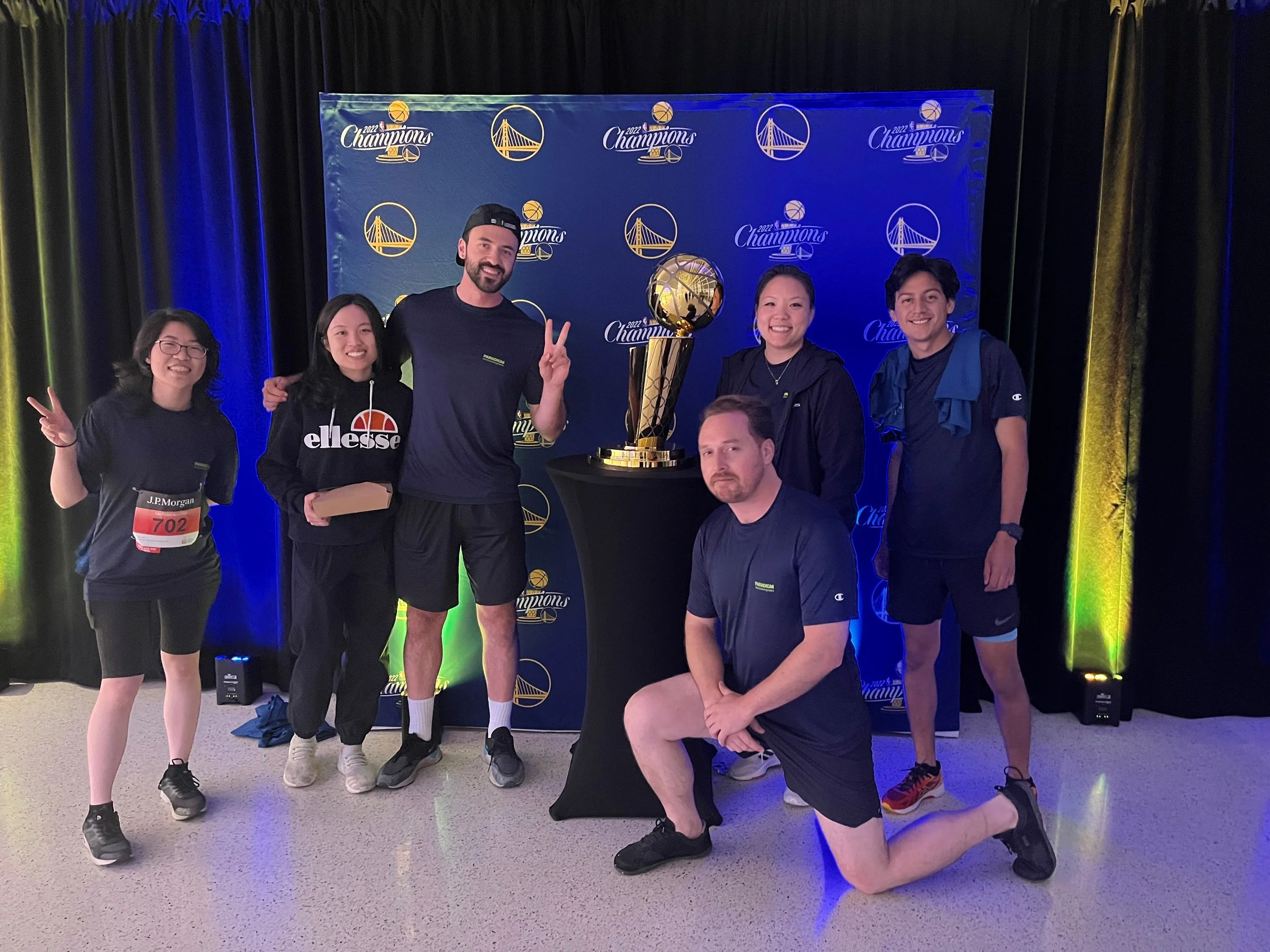 Group of six people posing in front of a backdrop with Golden State Warriors logos and a trophy. They are celebrating a victory, with one person showing a peace sign, and one kneeling in front of the group with a serious expression.