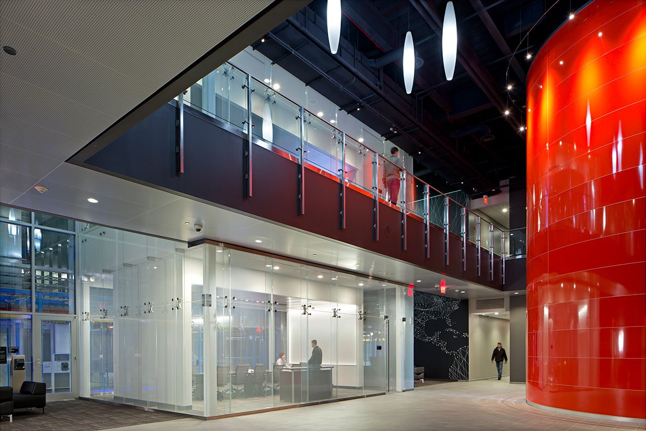 Modern office lobby with glass walls, a red cylindrical column, and a mezzanine level with glass railing. People are inside and walking through the space.