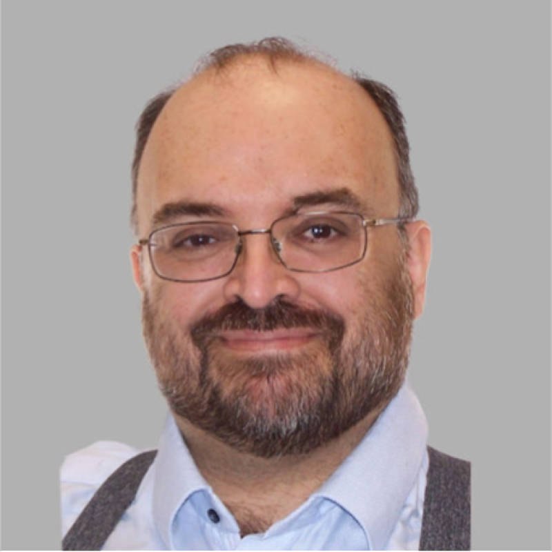 A middle-aged man with glasses, a beard, and a mustache smiling at the camera, wearing a light blue collared shirt and a dark vest, against a plain gray background.