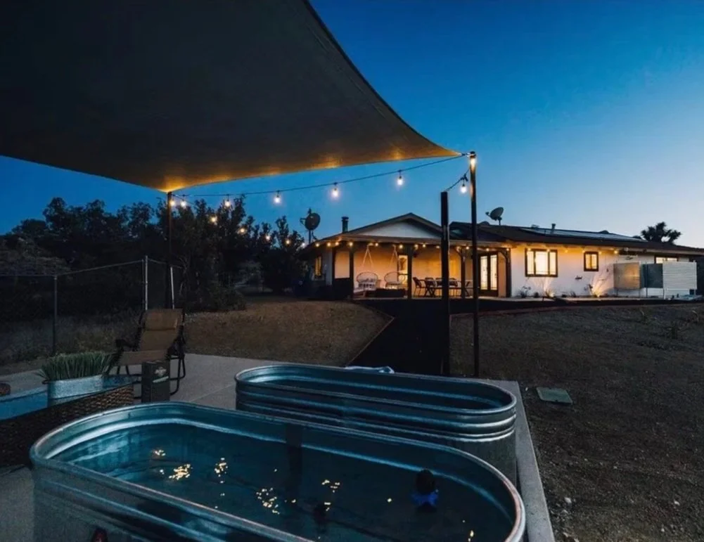 A backyard with a shade sail, outdoor furniture, and a hot tub, surrounded by desert landscape with Joshua trees and hills in the distance.