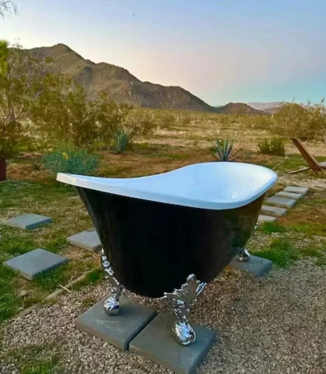 Bright kitchen with white walls, black cabinets, a wooden countertop, and a window with natural light. A kitchen sink with a black faucet, open shelves with dishes and decor, and a refrigerator are visible. In the background, there is a dining table with white chairs, a hanging light fixture, and a living area with a wall hanging and a door leading outside.