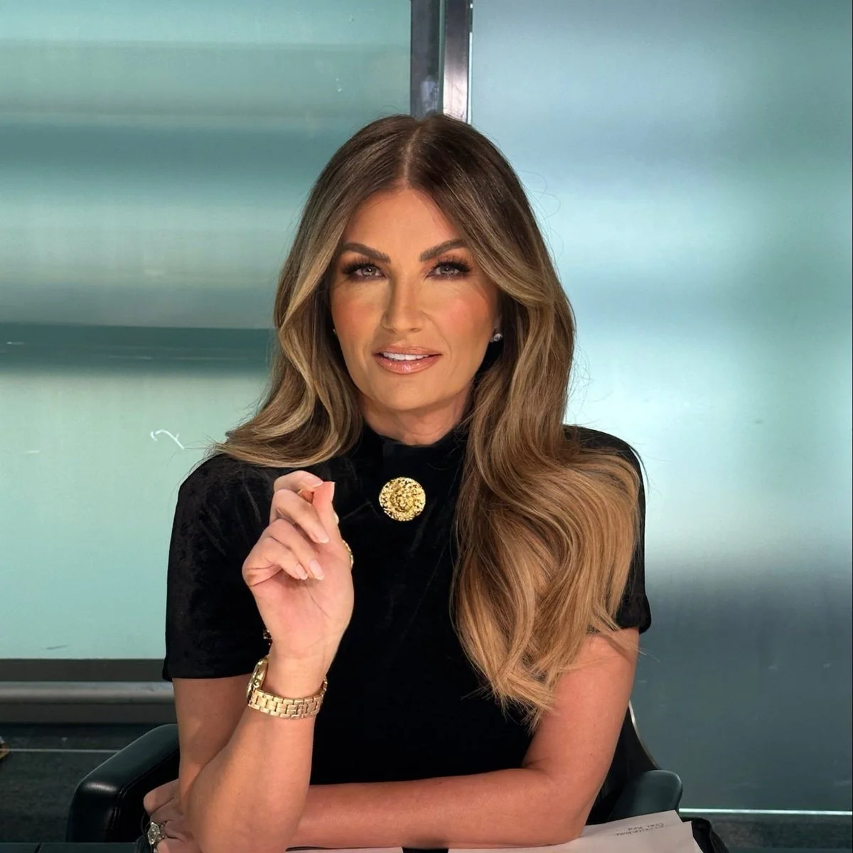 Karen Brady with long wavy hair wearing a black top with a gold brooch, sitting at a desk and holding a small object, in front of a frosted glass background.