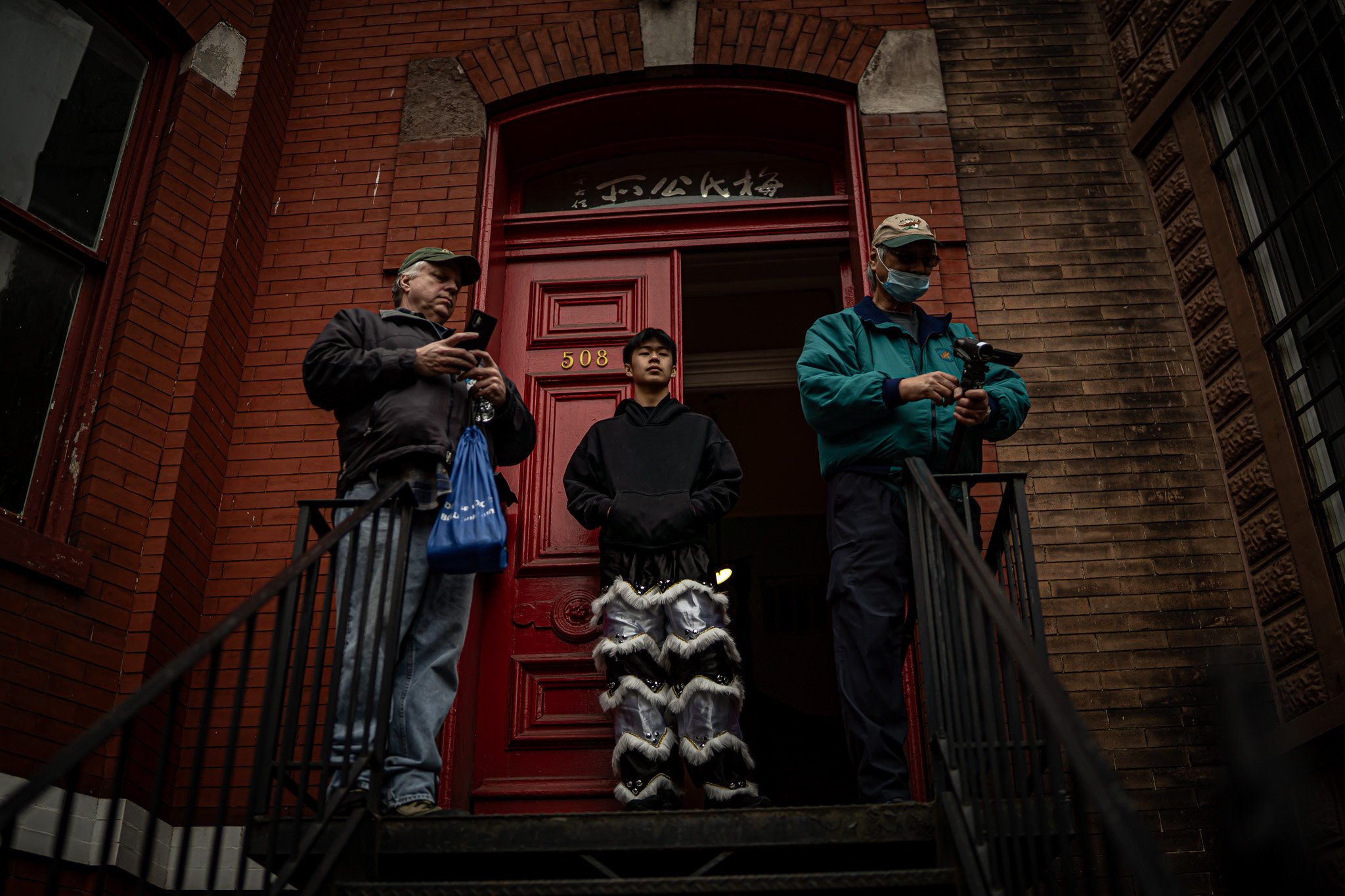 Three people standing outside a red brick building with the number 508 on the door. One man is looking at his phone, a young person dressed in a black hoodie and lion costume pants stands in the center, and another man is wearing a face mask and look