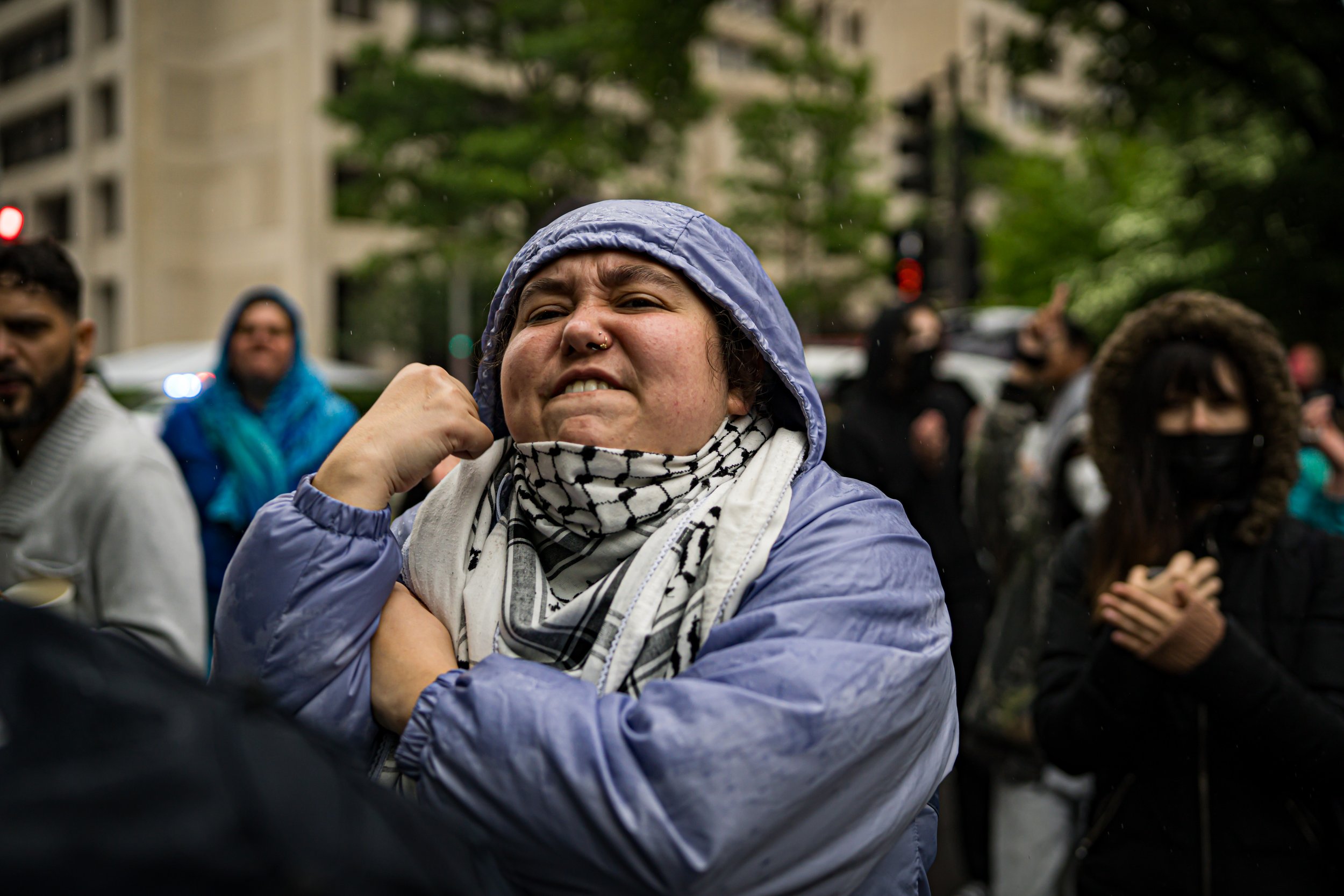 A woman in a blue raincoat and black and white scarf protests with a clenched fist and an angry expression, surrounded by other protesters in an urban setting.