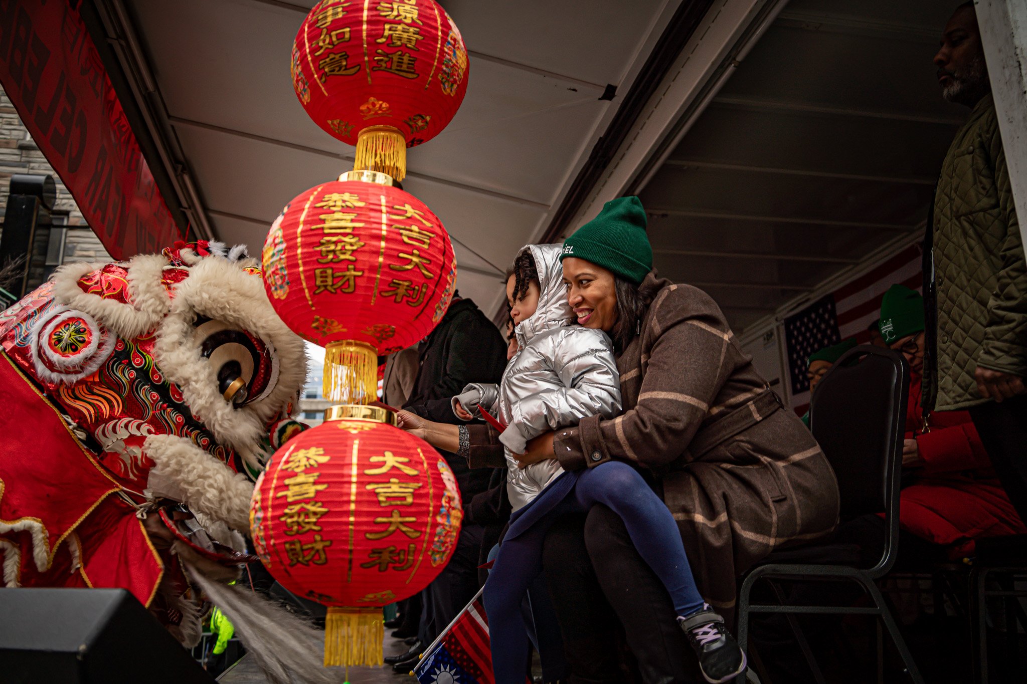 Woman and child at a Chinese New Year celebration, interacting with a lion costume performer beneath red lanterns with gold Chinese characters.