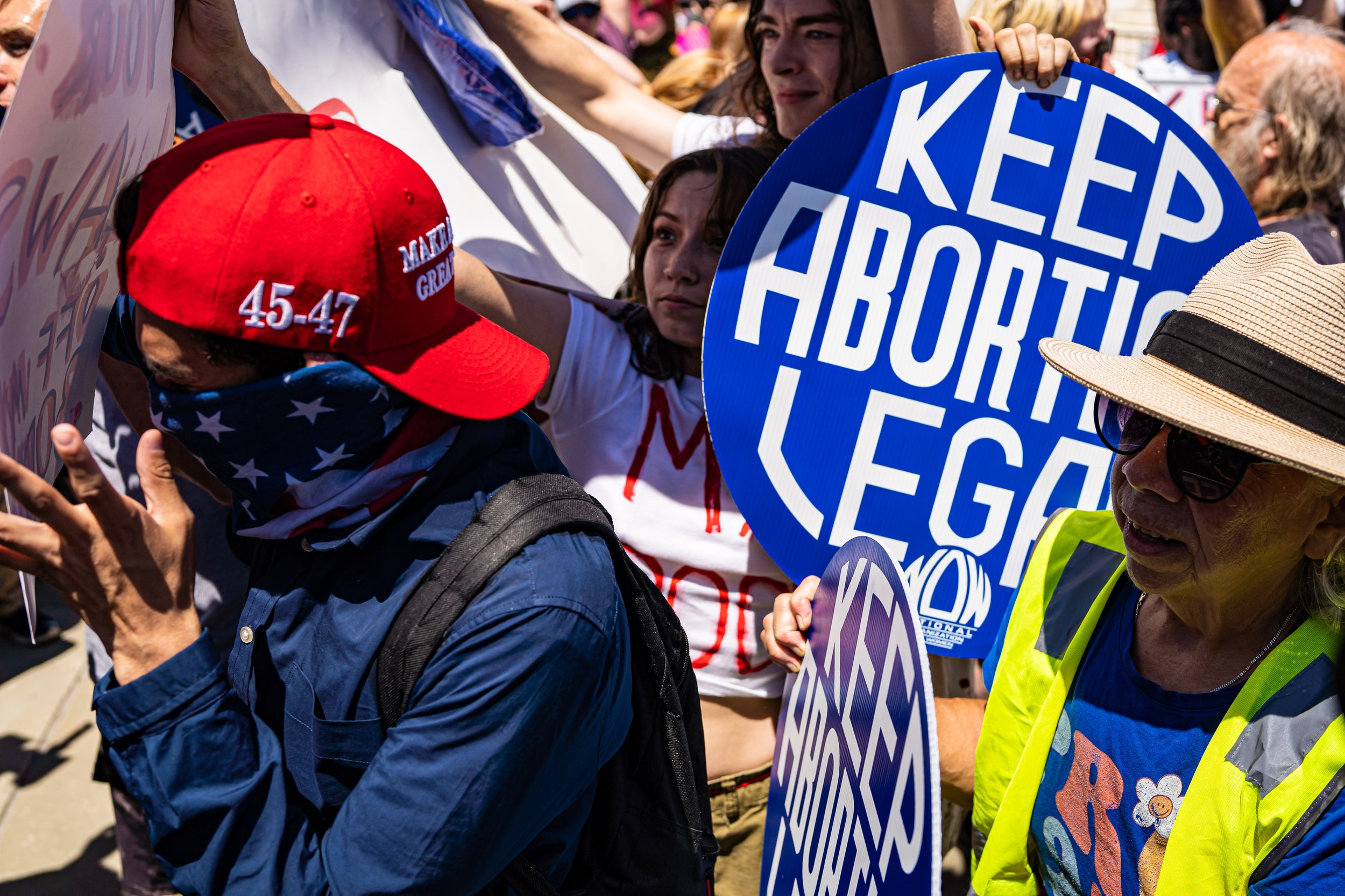 Protesters at an anti-abortion rally holding signs that say 'Keep Abortion Legal,' with one person wearing a red hat with white text and a person in sunglasses with a wide-brimmed hat.