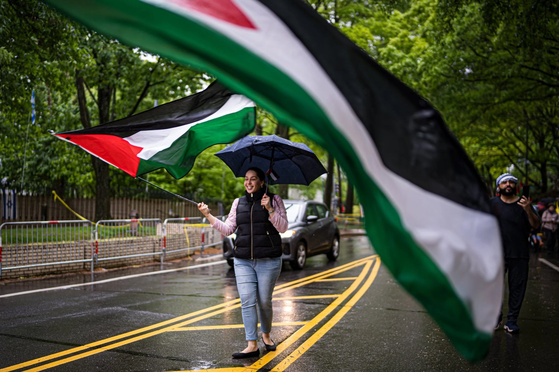 Woman holding an umbrella and waving a large Palestinian flag on a rainy street during a demonstration.