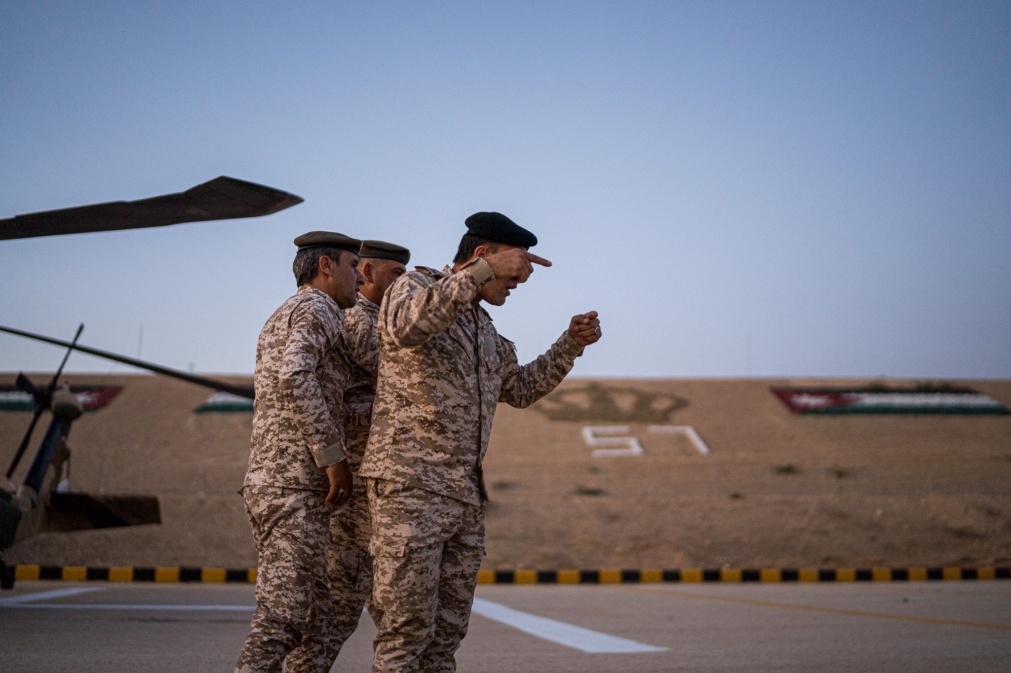 Three military personnel in camouflage uniforms standing outdoors, with one saluting, near military helicopters and a hill with the number 51 on it in the background