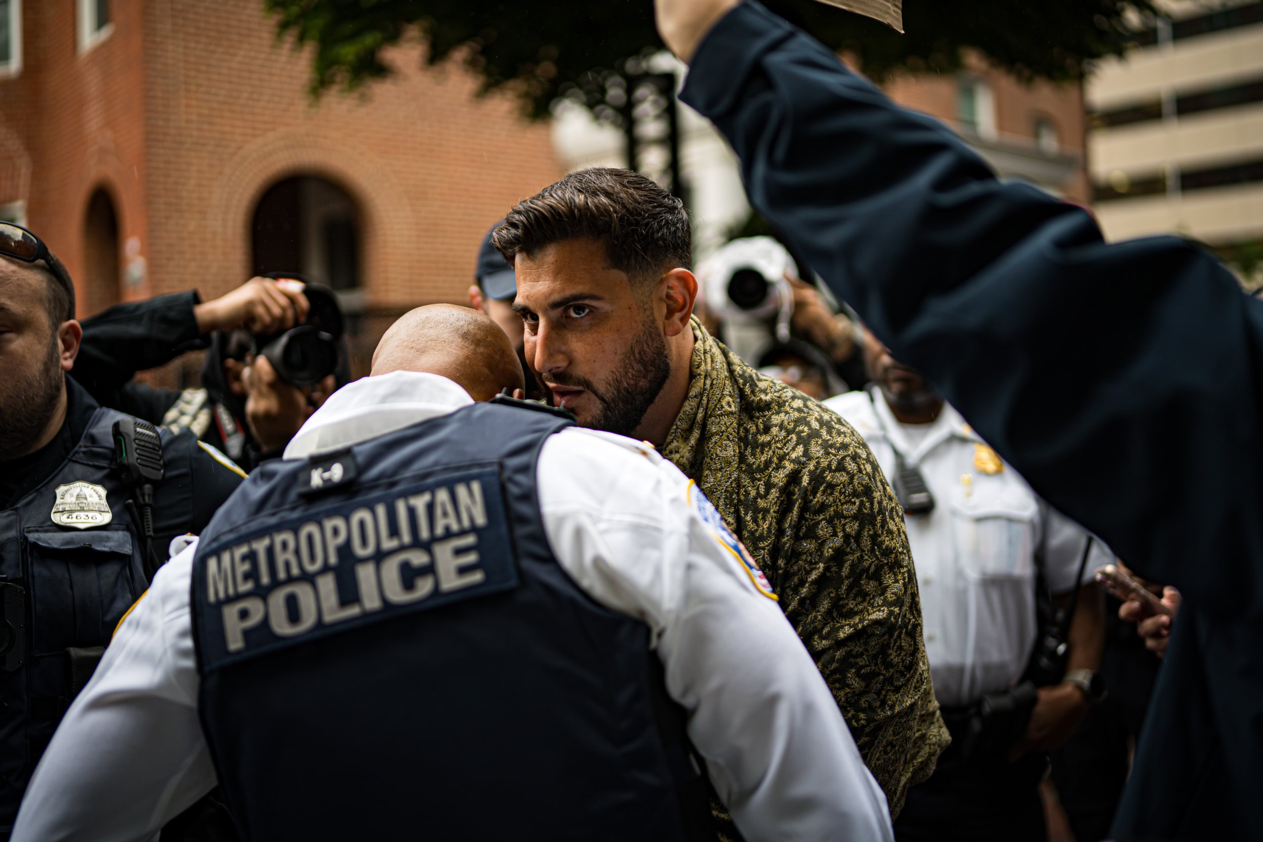 A man with a beard and patterned jacket confronts a group of police officers, including one in a Metropolitan Police vest, during an outdoor demonstration or protest, with other officers and photographers in the background.