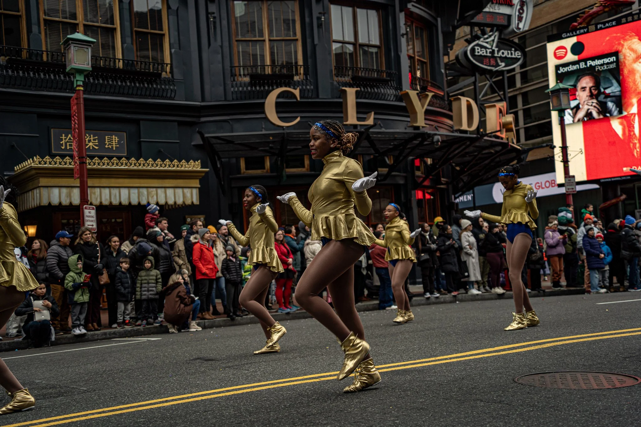 Group of dancers in gold costumes performing in a street parade, with a crowd of onlookers in front of a building labeled CLYDE and neon signs in the background.