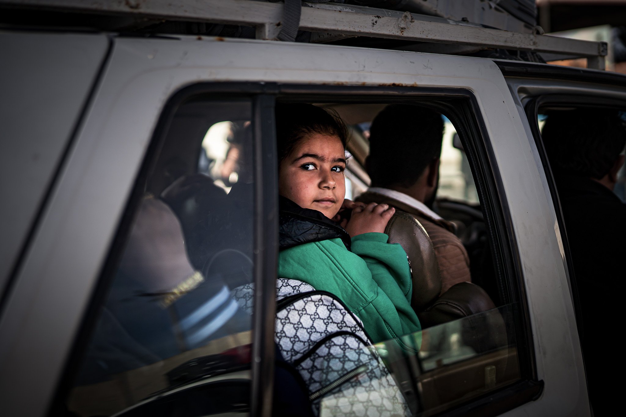 A young girl with dark hair and a green jacket looking out from an old vehicle's window, sitting with other people.