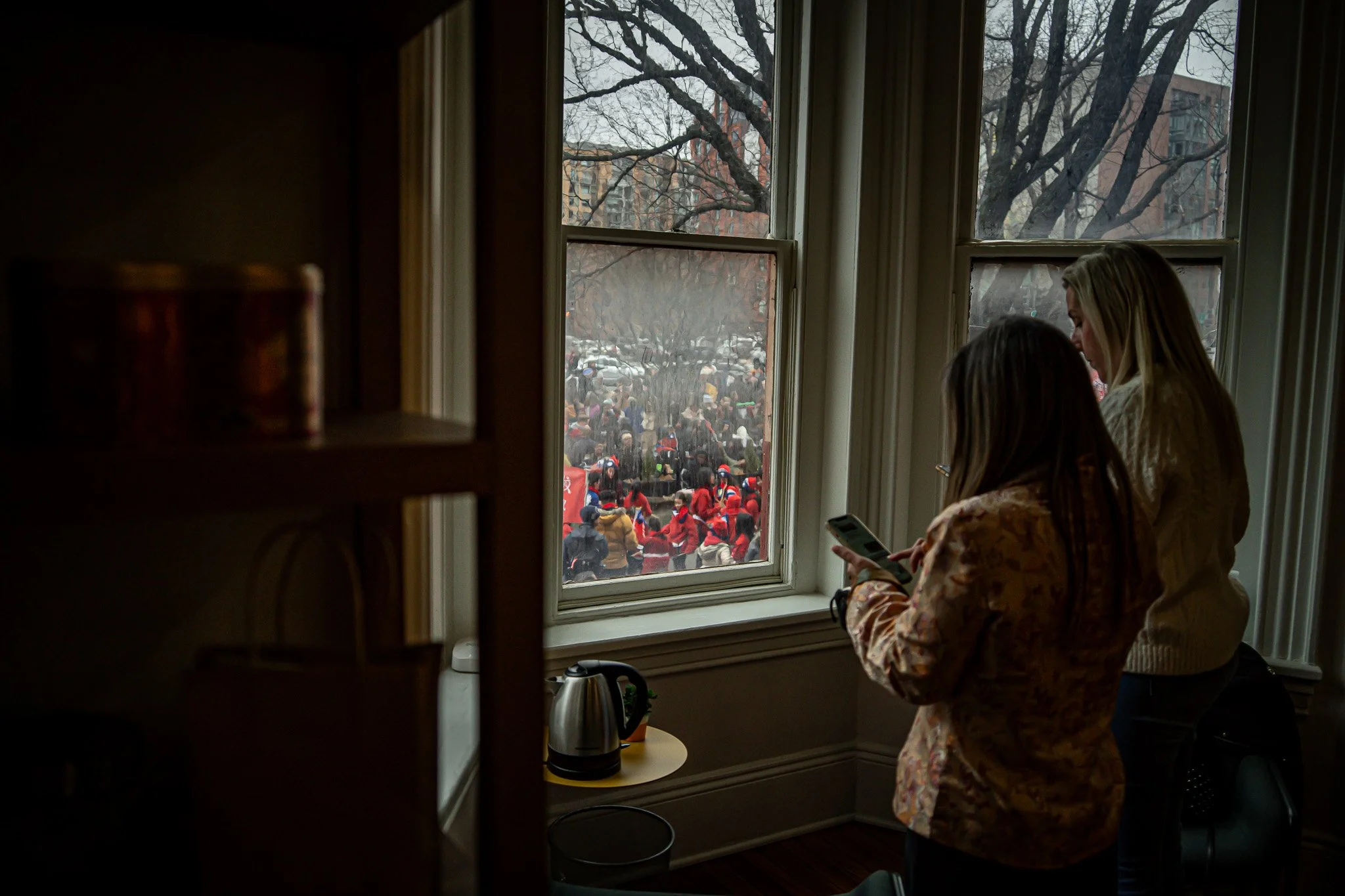 Two women standing inside a room by a window, observing a large crowd outside, with some wearing red and white hats.