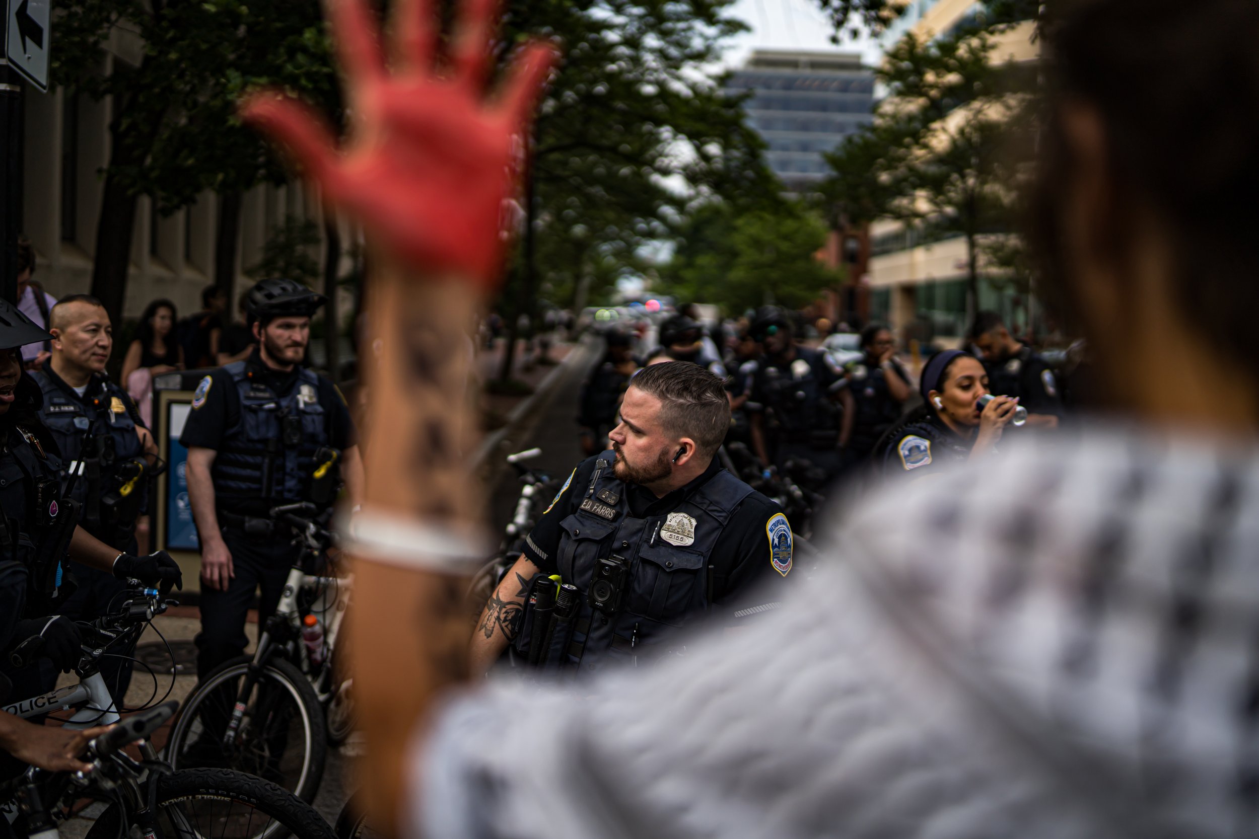A woman with a tattooed arm raising her hand dismissively in front of a group of police officers during a protest or demonstration on a city street.