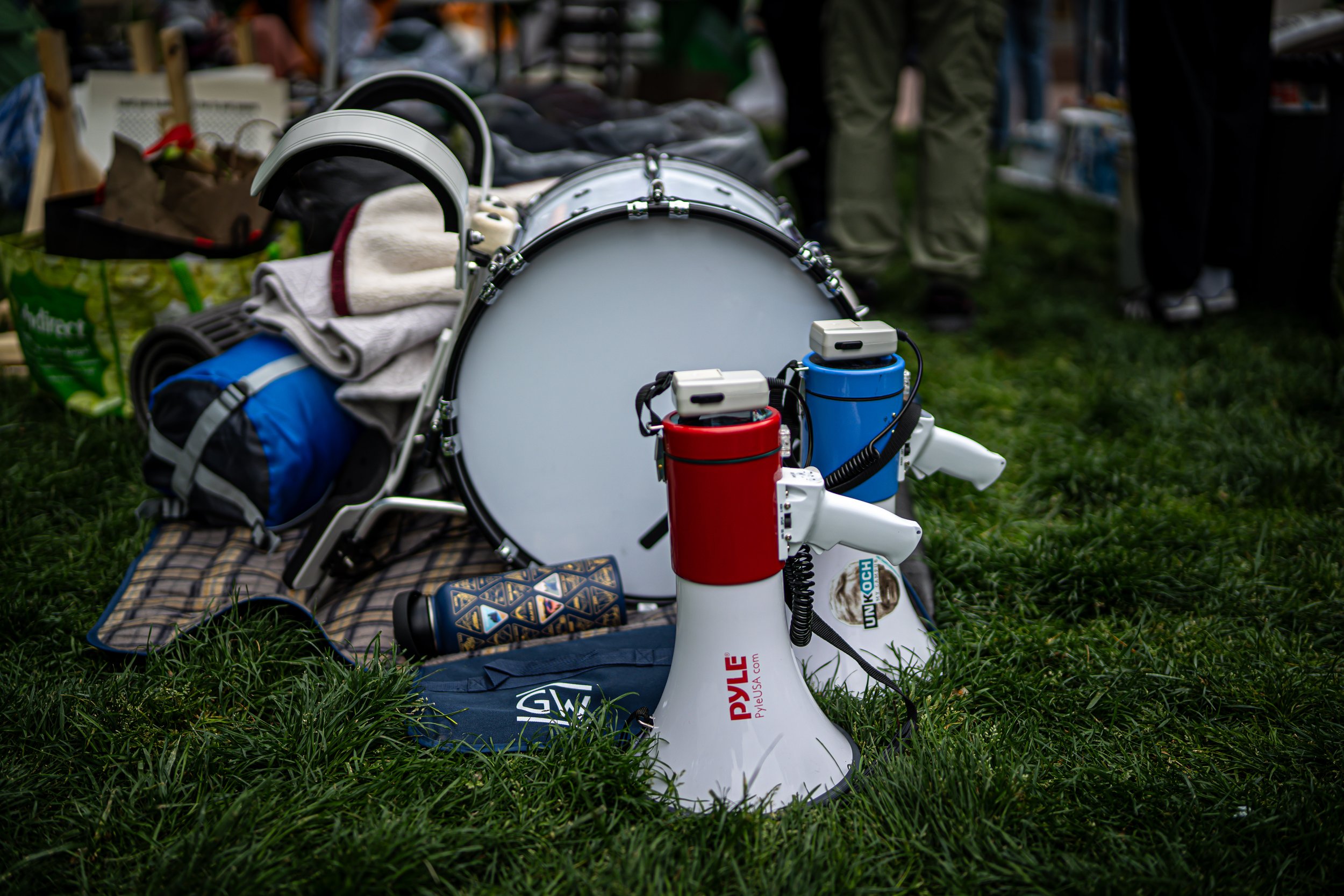A white megaphone with red and blue accents resting on green grass, surrounded by camping gear including a large drum, towels, and bags at an outdoor event.