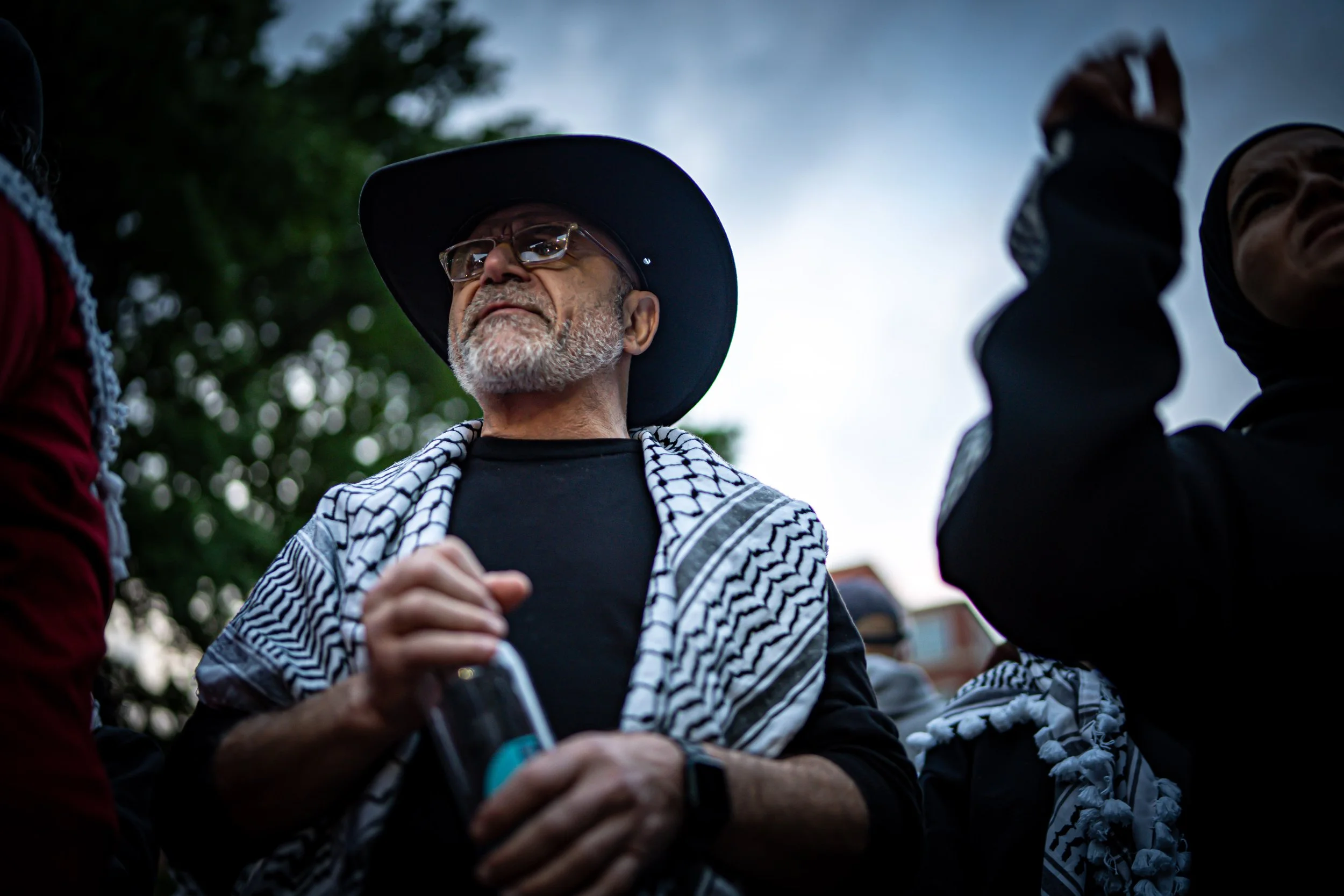 An elderly man with a beard, wearing glasses, a black wide-brimmed hat, and a keffiyeh scarf, stands among a group of people outdoors on a cloudy day.