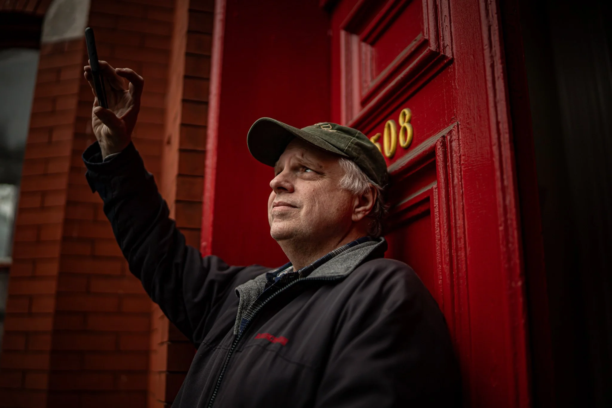 A middle-aged man in a dark jacket and baseball cap taking a selfie against a large red door with the number 208.