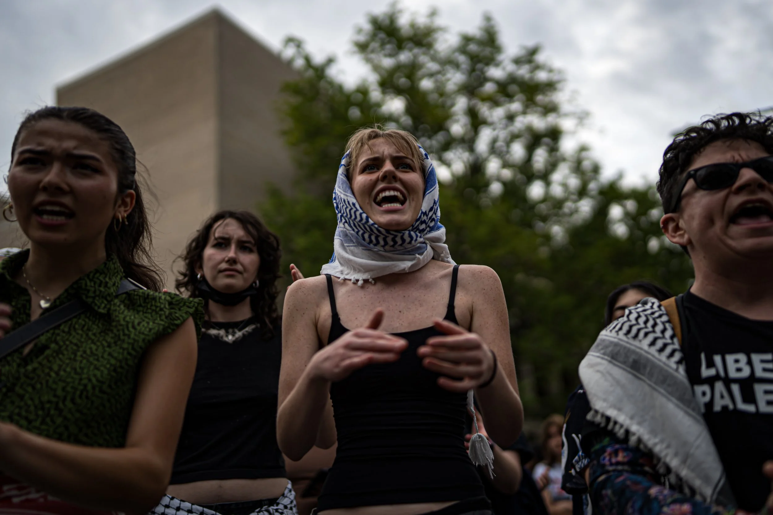Group of women protesting outdoors, looking distressed and angry, with trees and a building in the background.