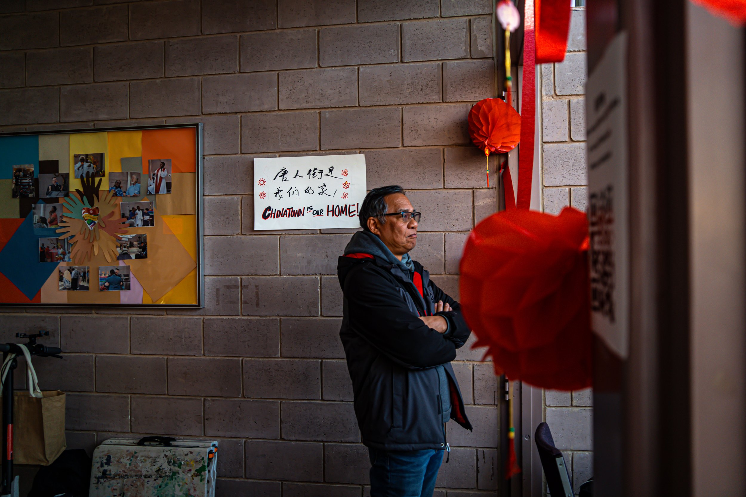 A man with glasses stands with arms crossed, looking at decorations on a wall inside a Chinatown community center. The wall has red paper lanterns and a sign with Chinese characters and the English words "Chinatown is our home!" next to a colorful ph