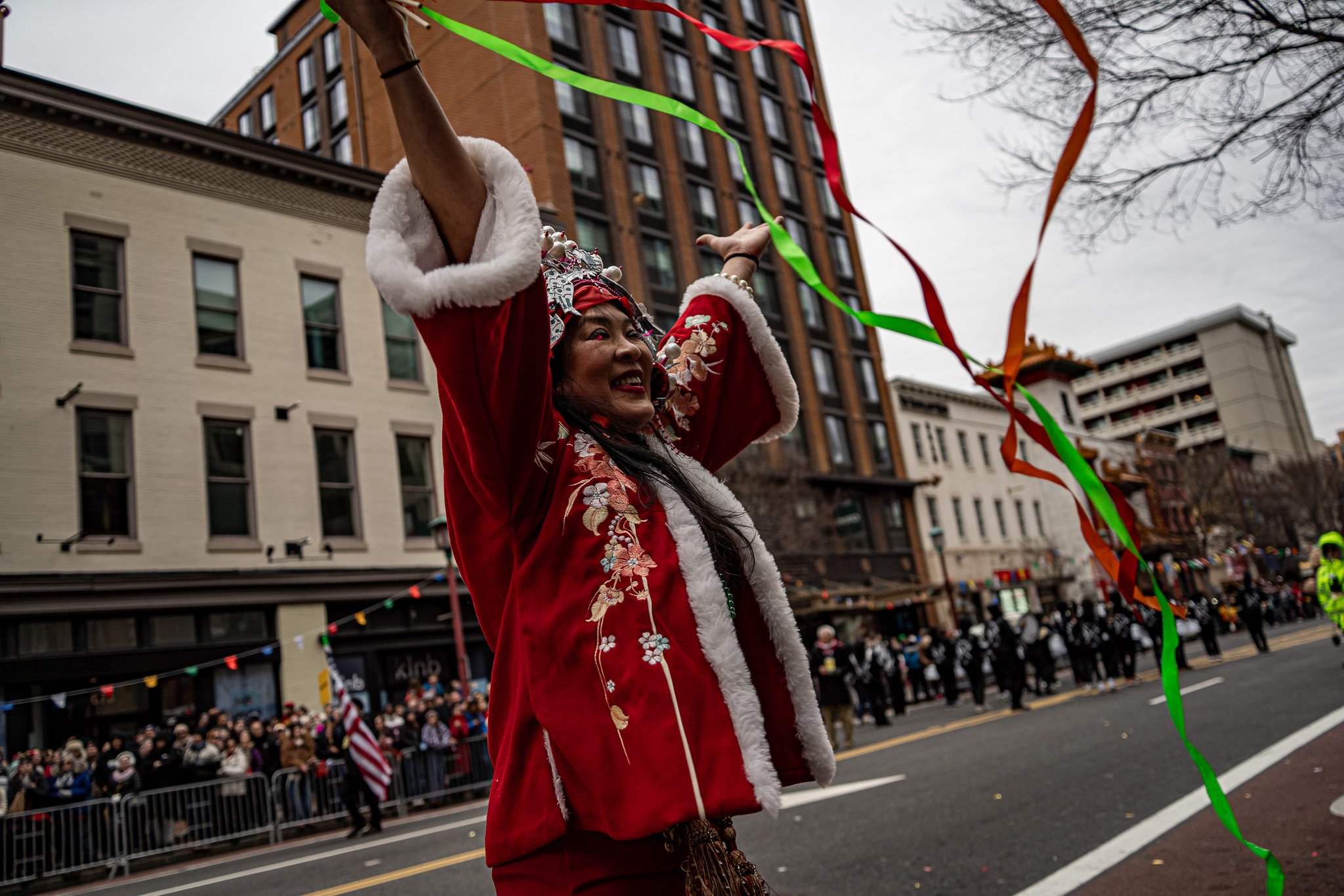 Woman dressed in traditional Chinese attire swinging colorful ribbons during a parade on a city street with spectators and buildings in the background.