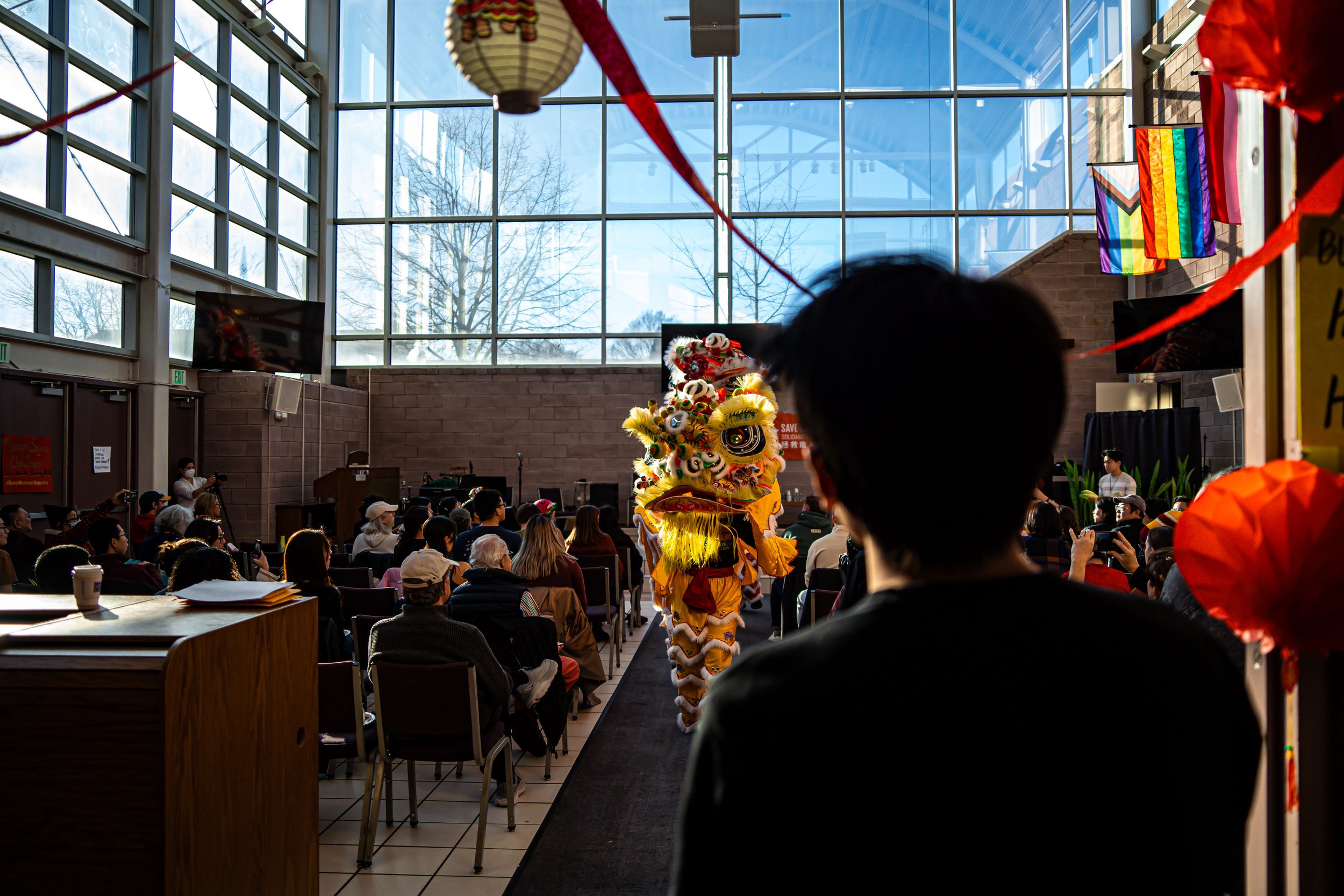 People attending a cultural event indoors with a lion dance performance in the center, during daytime with sunlight streaming through large glass windows.