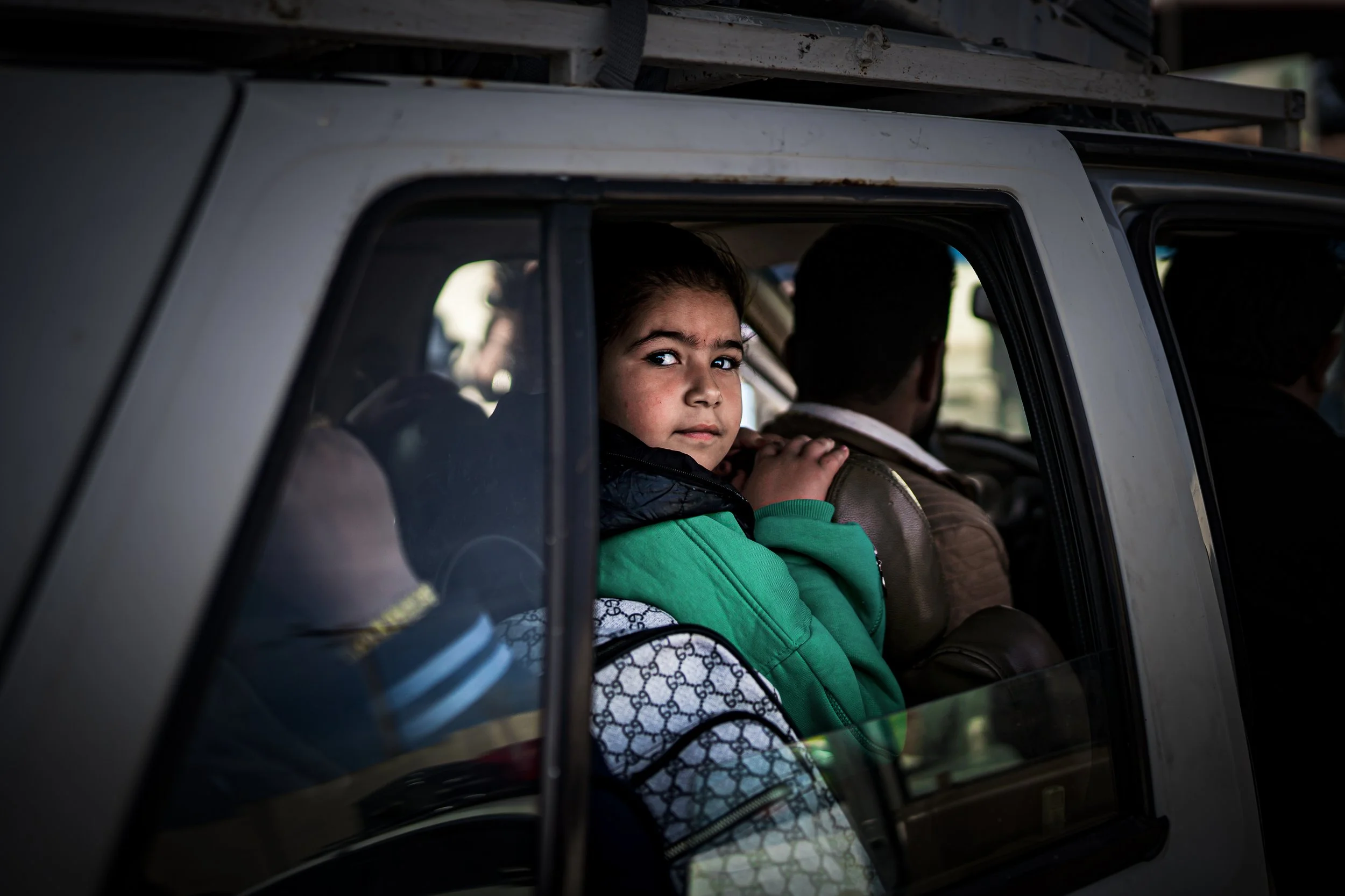 A young Syrian girl with a worried expression sitting in the backseat of a vehicle, looking out the window.