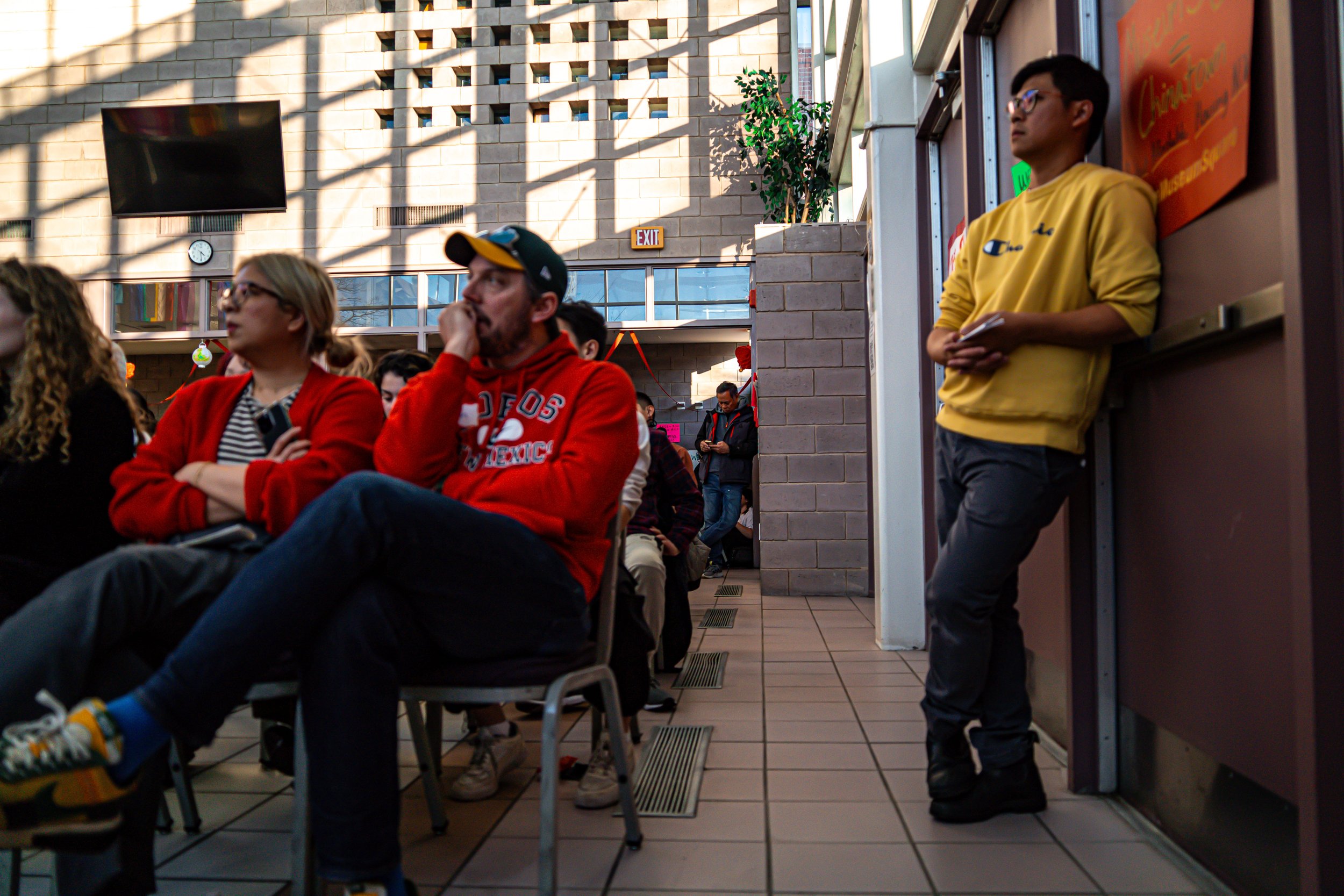 People seated in a room, some with arms crossed or resting their heads on their hands, listening attentively. A man in a yellow sweatshirt stands against the wall holding napkins or notes. The background shows a brick wall, a large flat-screen TV, an